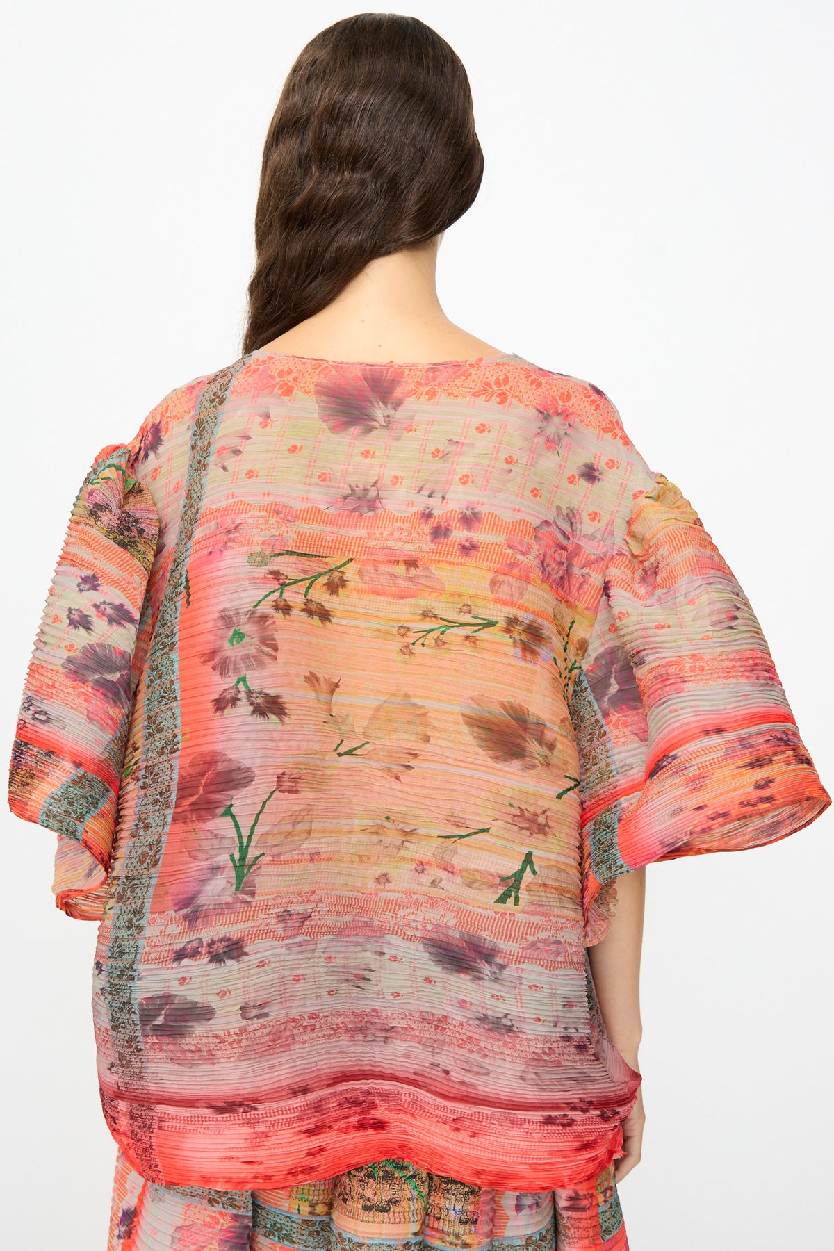 A person facing away, wearing the Julia Heuer Robin Handpleated Top in Bruno—an oversized, sheer, and loose-fitting sculptural piece crafted from recycled chiffon and adorned with floral and striped patterns in red, pink, and orange hues. Wavy brown hair drapes over one shoulder.