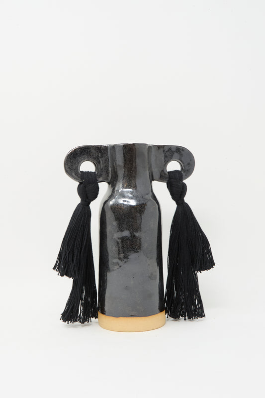 Karen Tinney’s Vase #606 in Black is a handmade black stoneware vase with a wide horizontal top and two large black tassels on each side, displayed against a plain white background.
