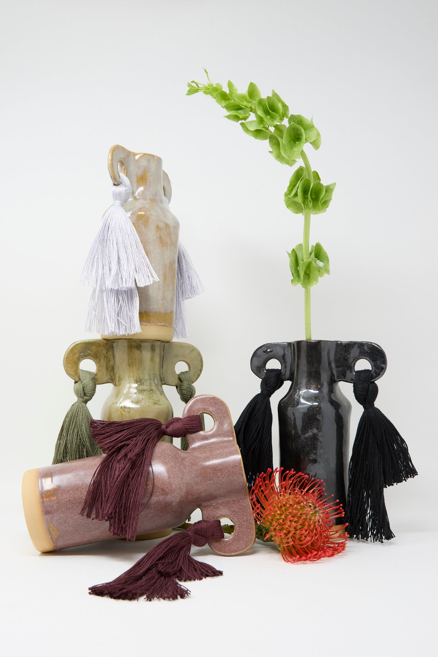 Three ceramic vases, including Karen Tinney's Vase #606 in Burgundy and a petite neutral-tone vase with large tassels, are arranged on a white surface. One displays a green stem and red-orange flower for an elegant effect.
