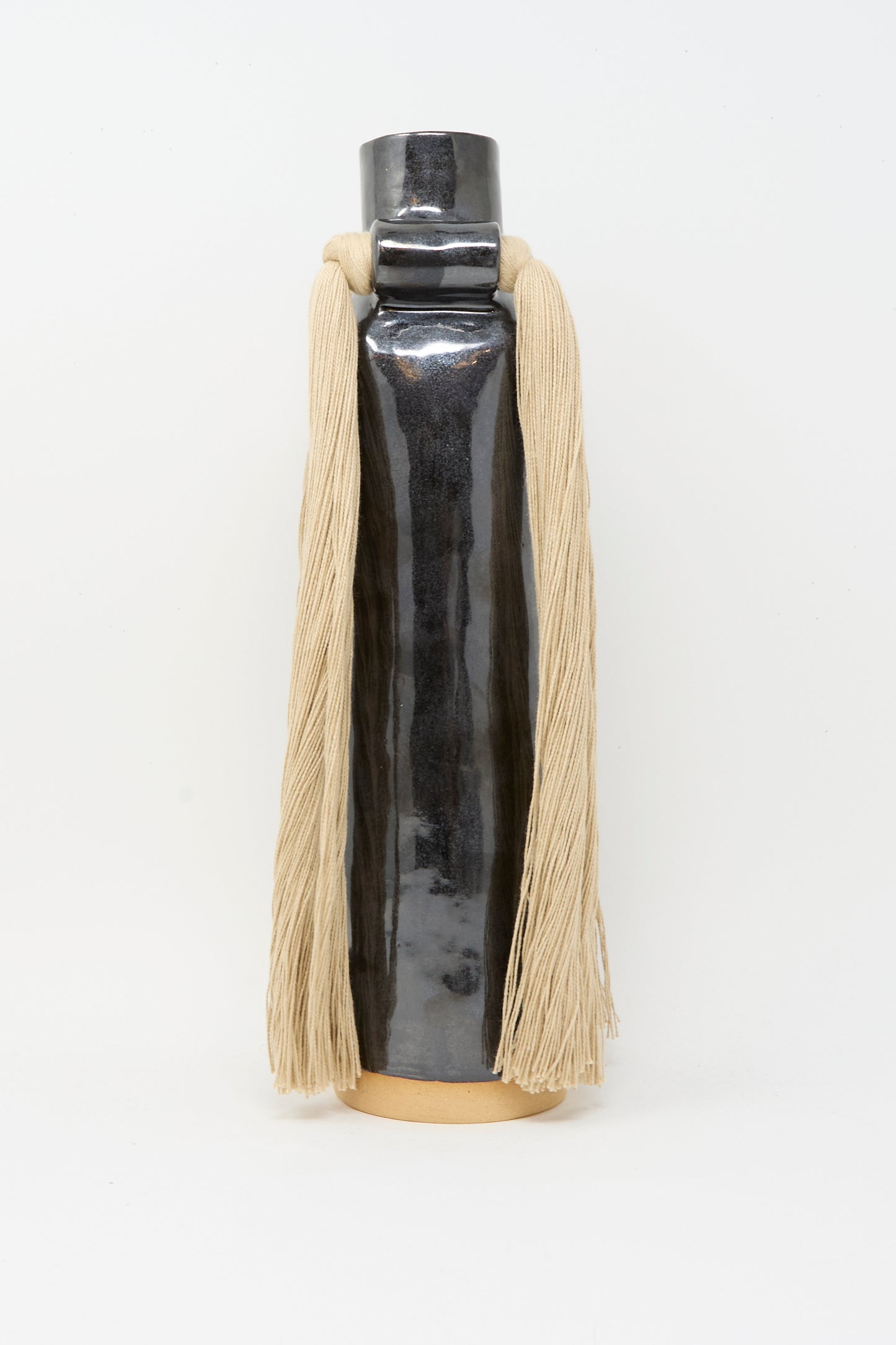 Vase #703 in Black by Karen Tinney is a tall, handmade stoneware vase with a glossy black glaze, cylindrical form, beige base, and long beige yarn elegantly draped from each side of the neck.