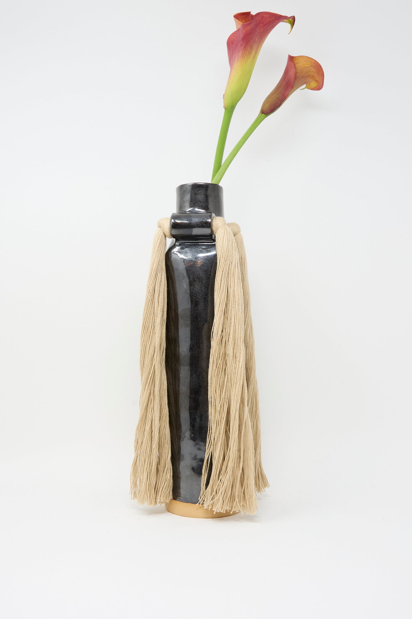 Karen Tinney's Vase #703 in Black, a tall glossy stoneware piece with beige tassels, holds two reddish-purple calla lilies against a white background.
