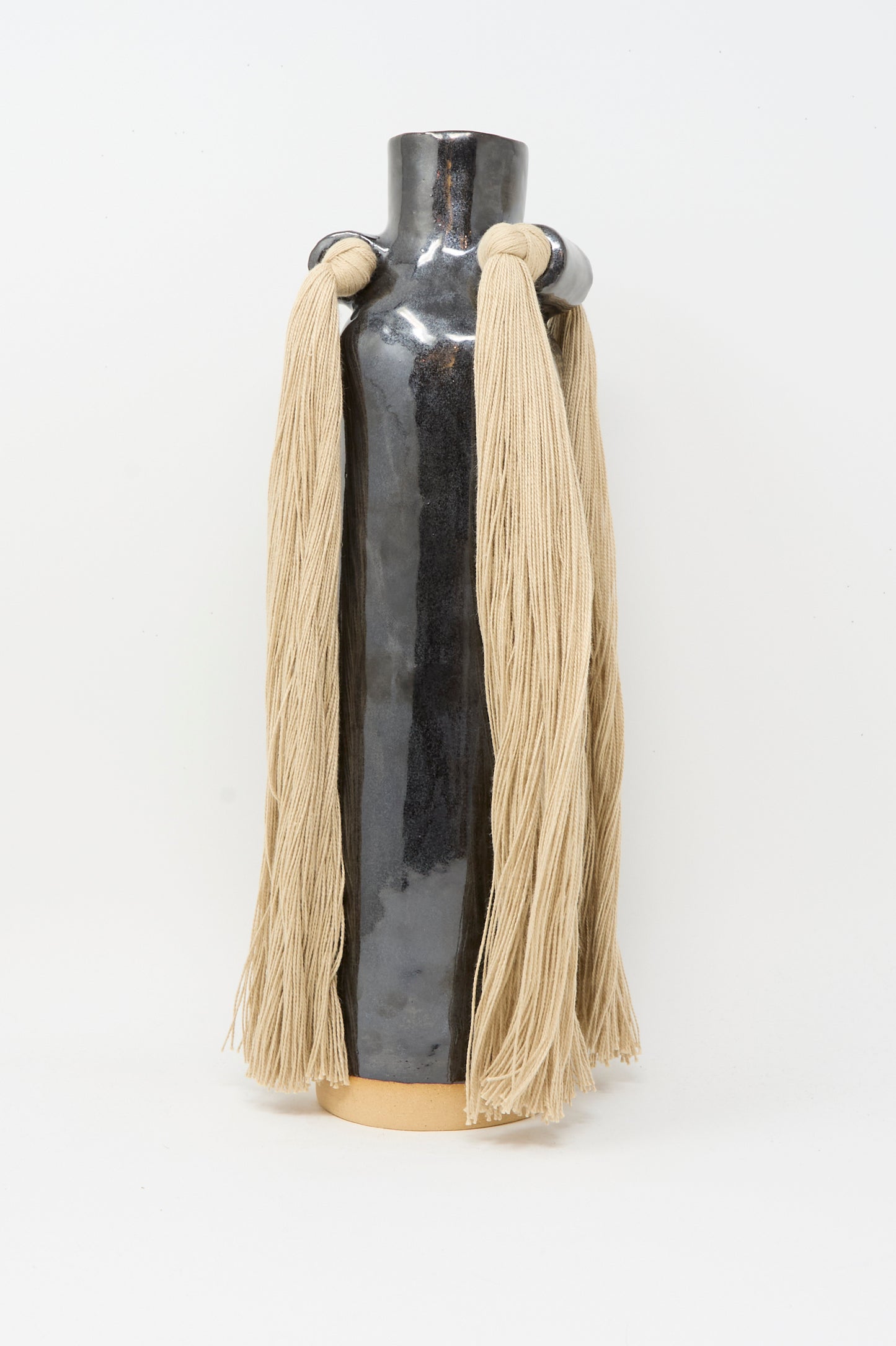 Karen Tinney's Vase #703 in Black is a tall, glossy black stoneware piece featuring two circular handles adorned with long beige yarn strands, standing on a light base against a white background.