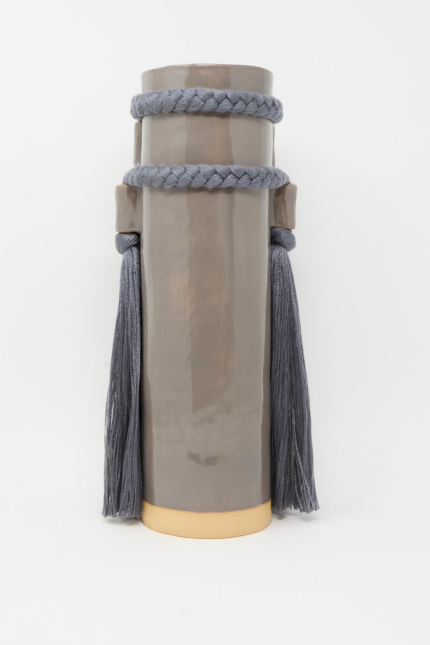 Karen Tinney's Vase #735 in Charcoal is a tall cylindrical handmade stoneware vase featuring a glossy taupe finish, braided rope with long tassels near the top, and a gold-colored base, shown against a white background.