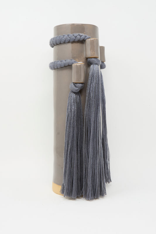 Karen Tinney’s Vase #735 in Charcoal is a tall, cylindrical ceramic piece featuring two braided cords and long gray tassels around the top, inspired by Vermont pottery.
