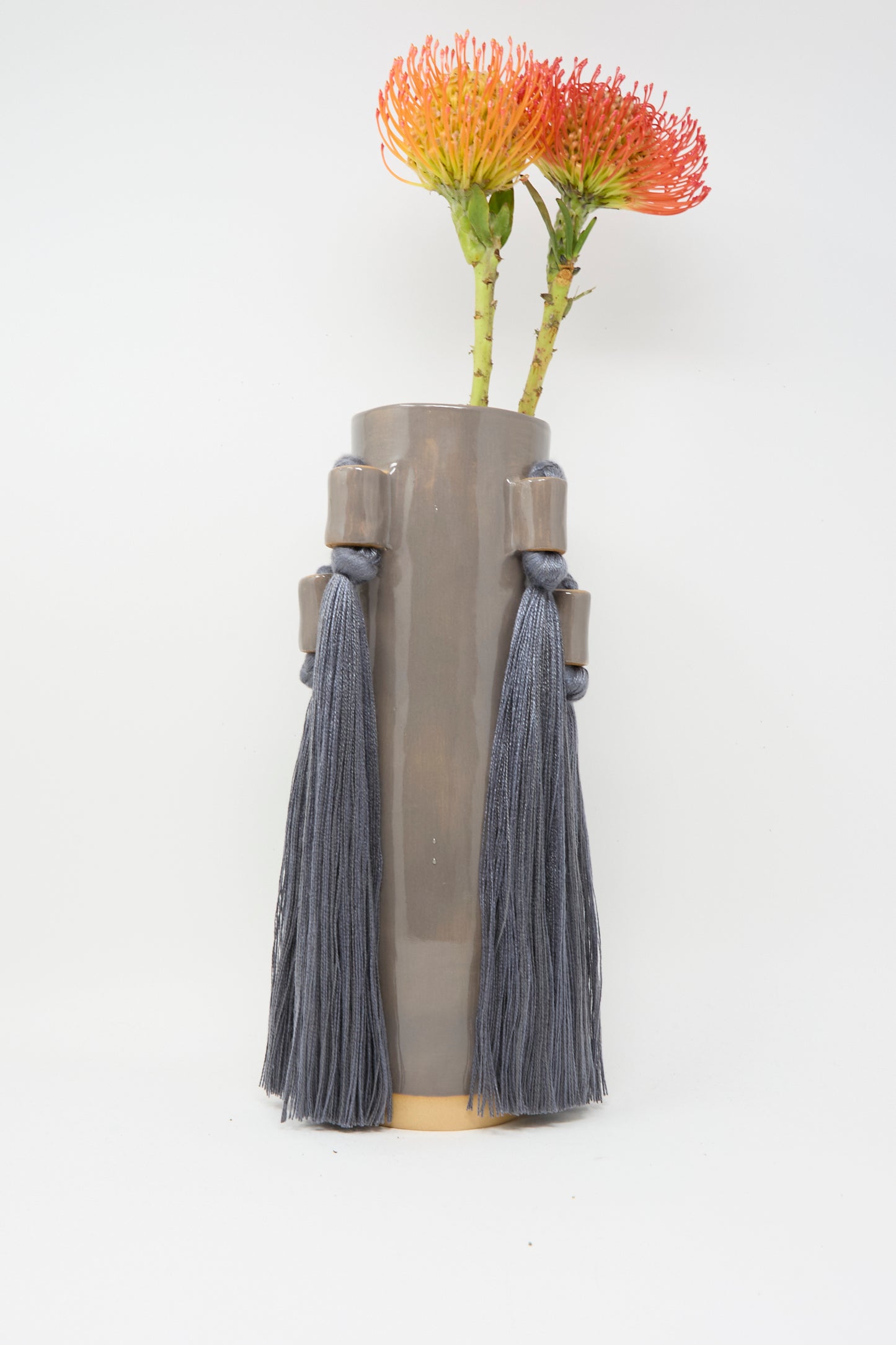 Karen Tinney’s Vase #735 in Charcoal, a handmade stoneware piece with two large blue tassels, is shown holding orange pincushion protea flowers against a white background.