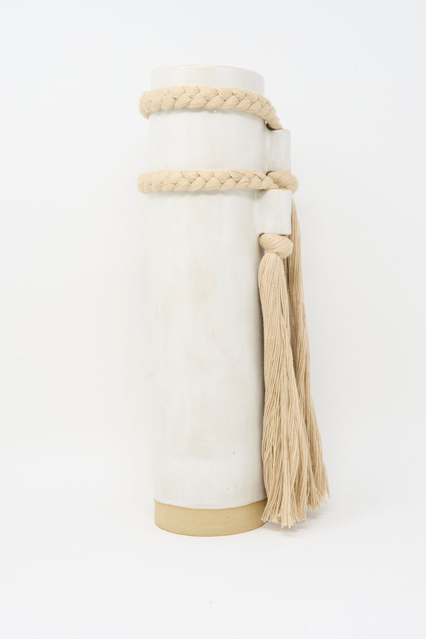 Karen Tinney’s Vase #735 in White is a tall, handmade cylindrical vase with a beige base, finished in white glaze and wrapped with a thick braided rope accented by two long tassels on the right.