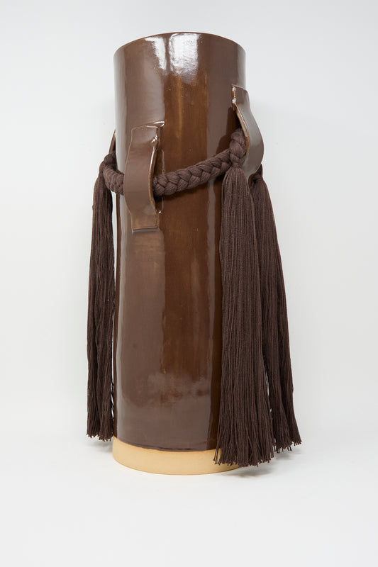 Handmade in Vermont, the Karen Tinney Vase #800 in Brown is a tall cylindrical stoneware vase with two handles, featuring a braided dark brown rope and tassels. Shown here against a plain background.