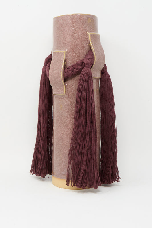 Vase #800 in Burgundy by Karen Tinney is a tall, cylindrical pink stoneware vase with two rectangular handles, featuring a braided burgundy cord and long tassels—a unique piece inspired by Vermont pottery.