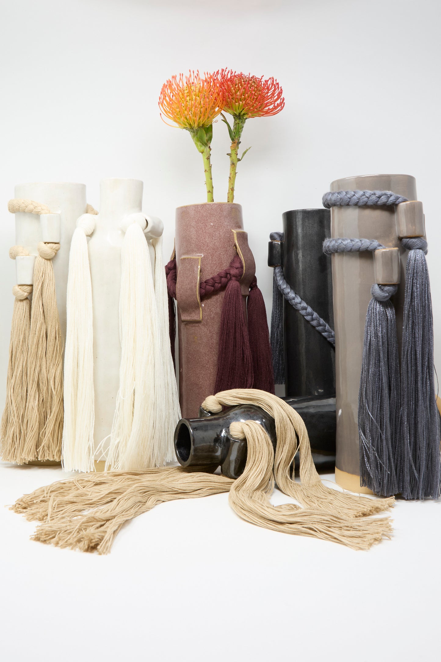Several Karen Tinney stoneware vases in neutral tones with long tassels are displayed together; Vase #800 in Burgundy, handmade, holds two orange protea flowers, adding a touch of Vermont pottery charm.