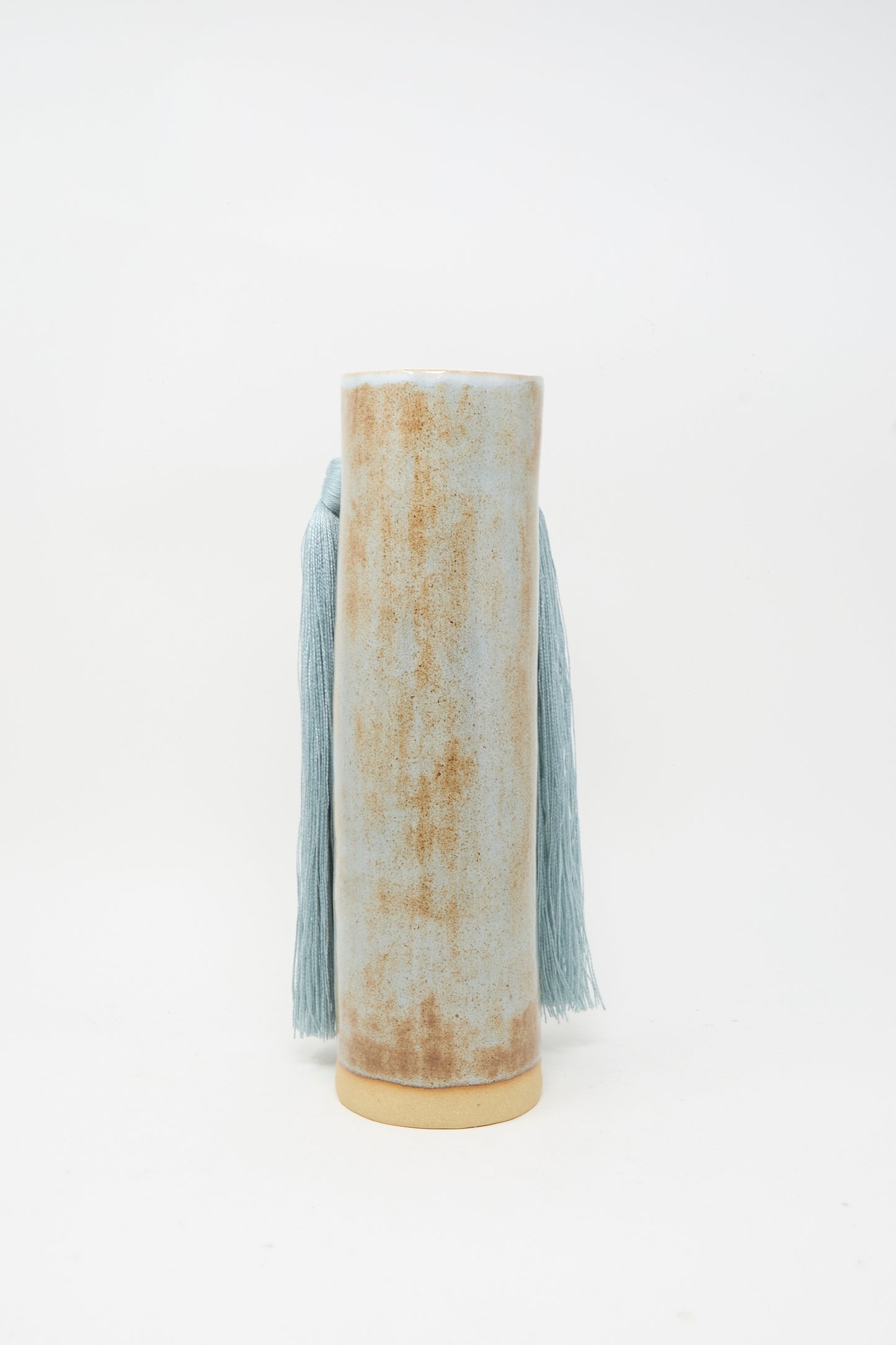 Vase #531 in Blue by Karen Tinney is a tall, cylindrical handmade stoneware vase with a distressed beige and light blue finish. It features long blue tasseled cords hanging from the sides and is set against a plain white background.