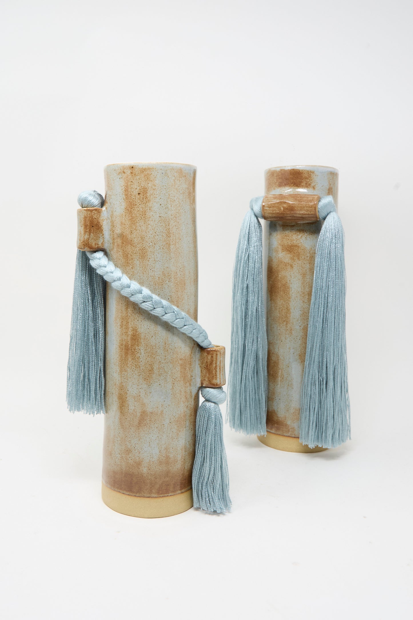 Karen Tinney’s Vase #695 in Blue is a tall, cylindrical stoneware vase with a brown and beige finish, elegantly adorned with light blue braided rope and long tassels for a refined handmade touch.