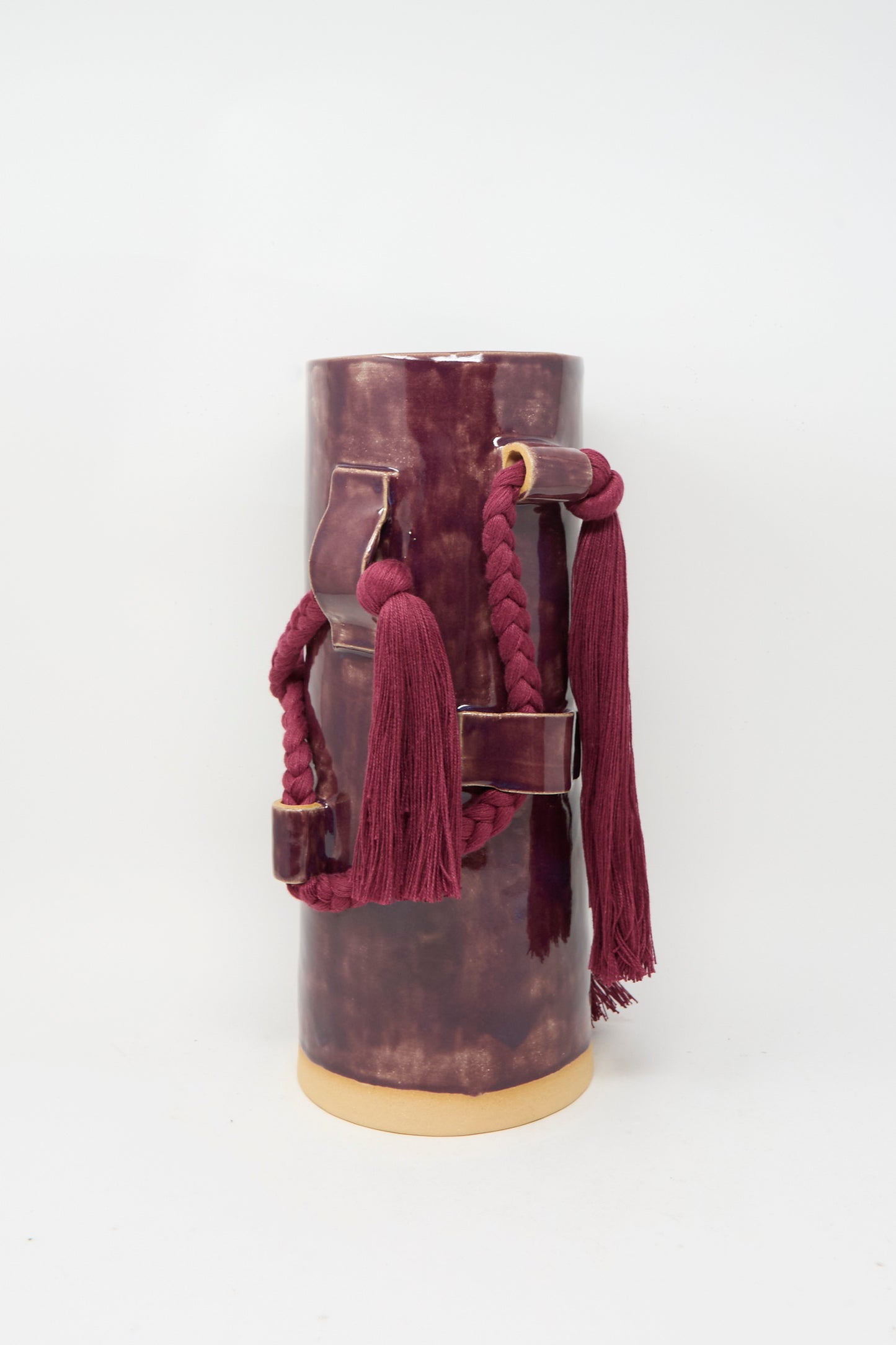 Karen Tinney’s Vase #846 in Plum, a tall cylindrical stoneware piece with braided cord, tassels, and leather strap accents, stands on a light background.