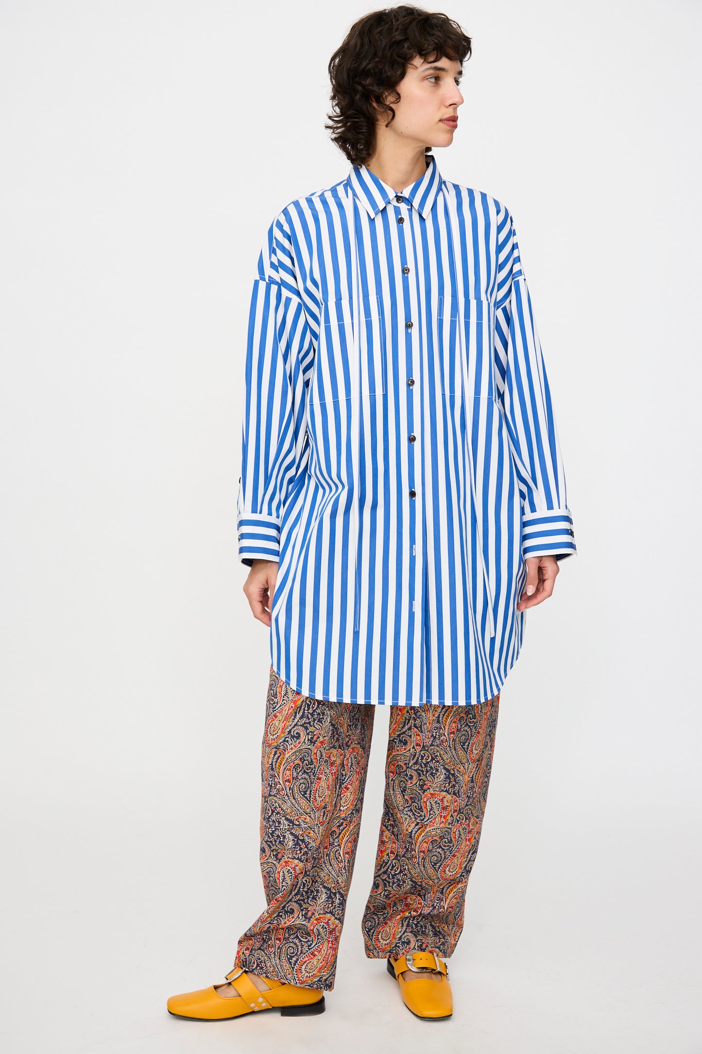 Model wears the KasMaria Cotton Poplin Oversized Dress with Tie in Stripe, paired with paisley print wide-leg pants and yellow shoes, set against a plain white background.