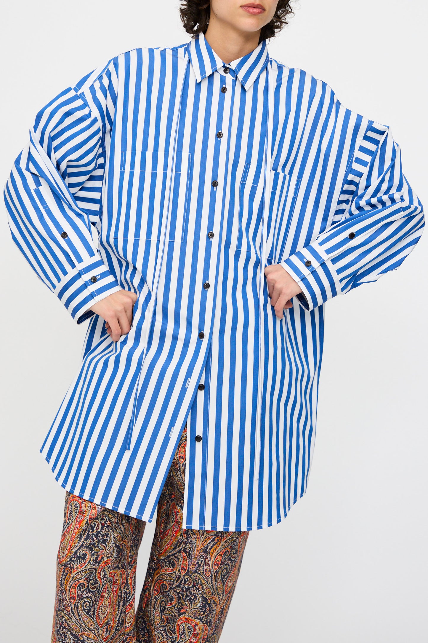 A person wears the KasMaria Cotton Poplin Oversized Dress with Tie in Stripe, featuring blue and white stripes, against a plain background.