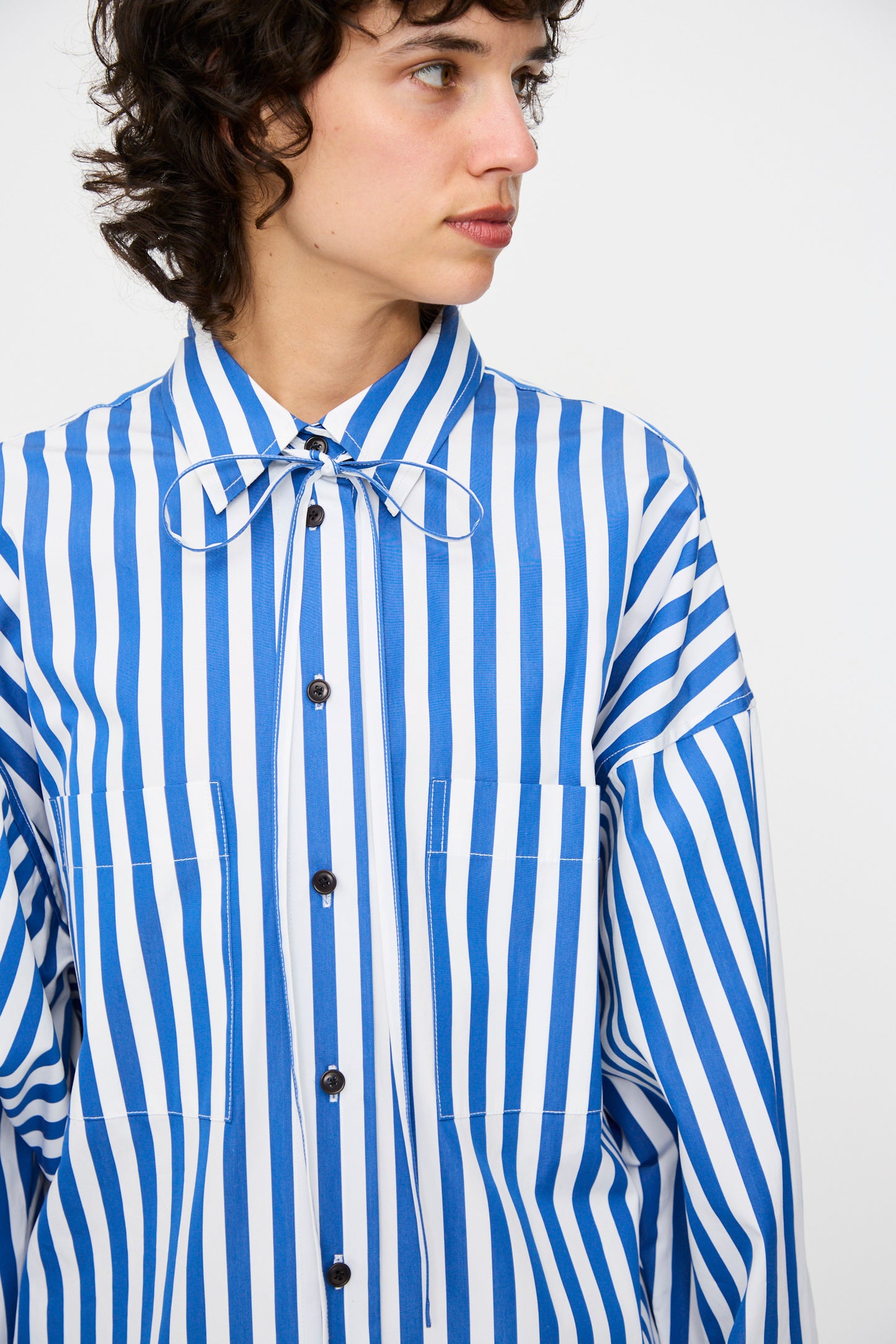 A person wearing the KasMaria Cotton Poplin Oversized Dress with Tie in Stripe, featuring blue and white vertical stripes, a bow at the collar, front pockets, and an oversized fit, looking to the side.
