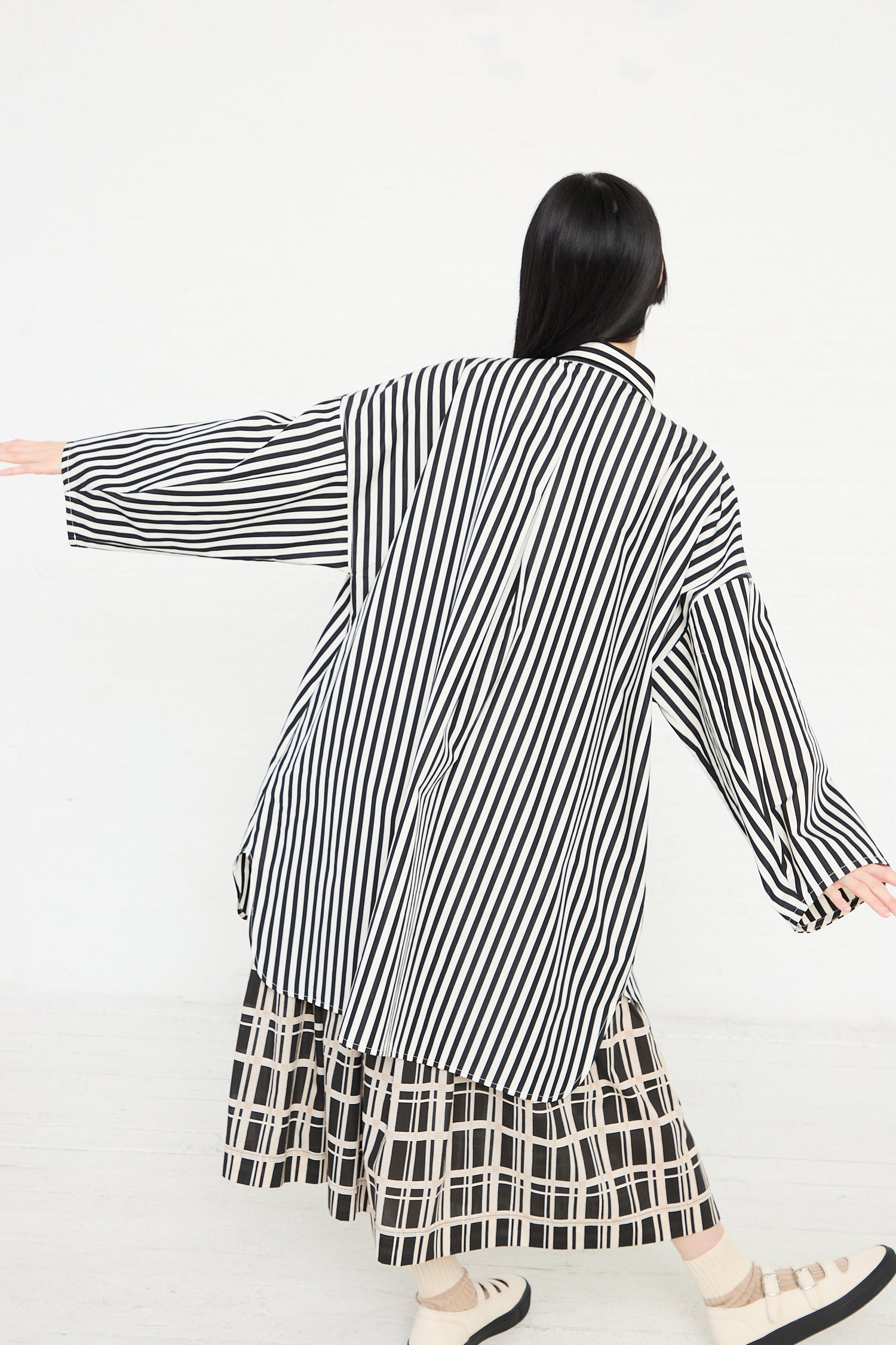 Woman posing in a KasMaria Cotton Poplin Oversized Smock Dress in Stripe and patterned skirt with her back to the camera and arms extended.
