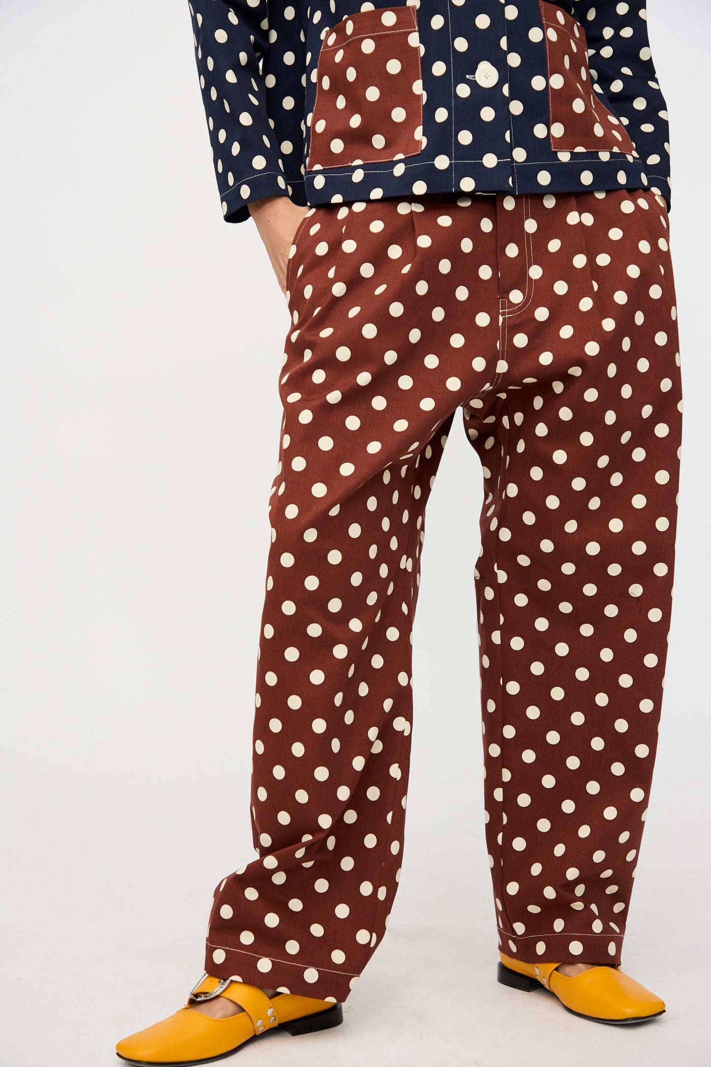 A person models KasMaria’s Cotton Twill Pleated Trouser in Polka Dot, paired with a navy polka dot jacket and yellow shoes, standing against a plain background. Made in NYC.