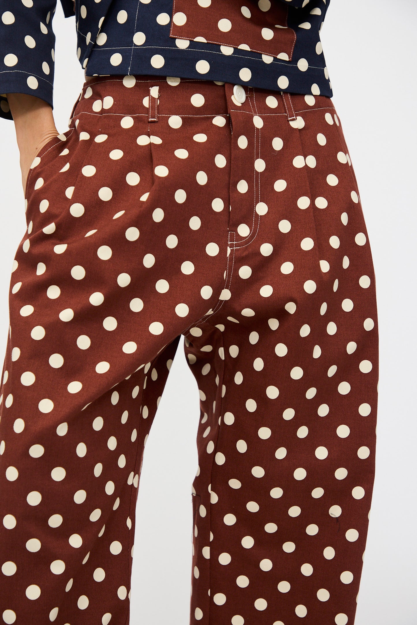 Model wearing KasMaria’s Cotton Twill Pleated Trouser in Polka Dot—brown cotton twill pants with white polka dots, paired with a navy top. Shown waist to mid-thigh, one hand casually placed in pocket. Made in NYC.