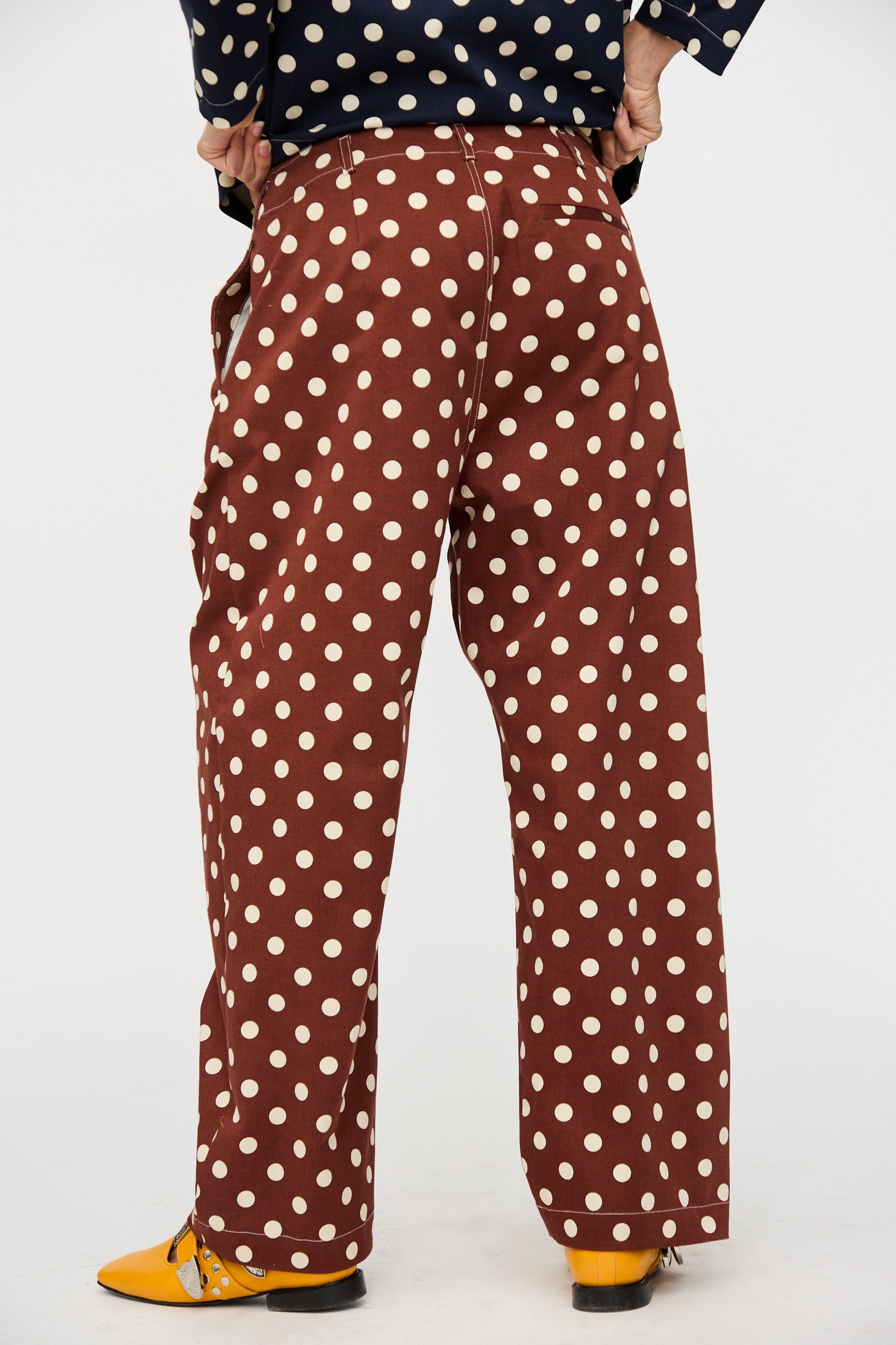 A person stands away with hands on hips, wearing KasMaria’s Cotton Twill Pleated Trouser in Polka Dot, a navy top, and yellow shoes.