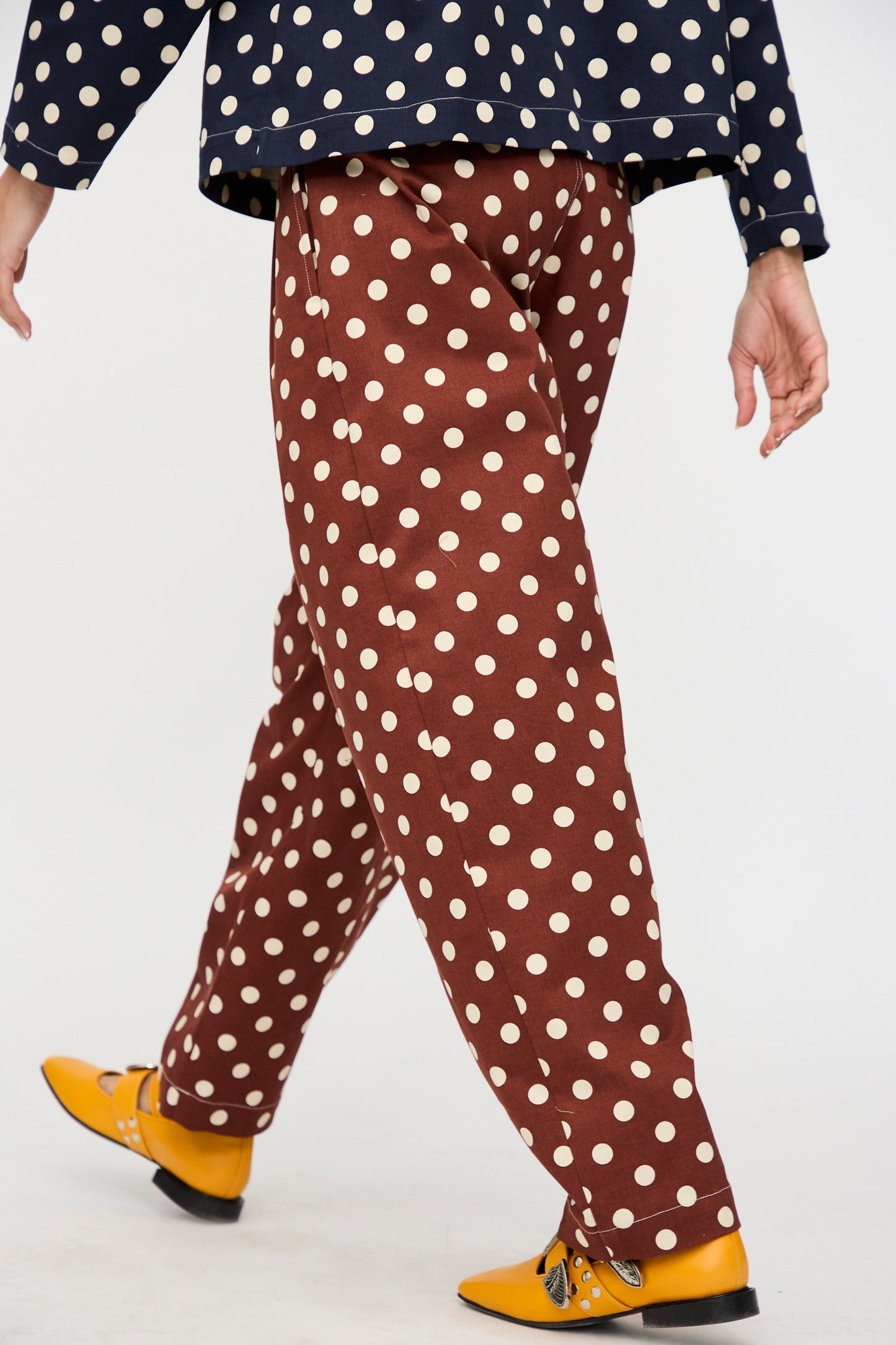 Someone walks against a plain background wearing KasMaria's Cotton Twill Pleated Trouser in Polka Dot, paired with a navy polka dot top and yellow shoes.