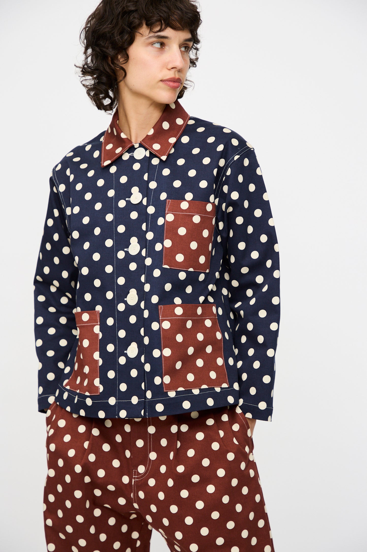 A person wears the KasMaria Cotton Twill Shirt Jacket in Polka Dot with contrasting brown and white patch pockets, paired with matching brown polka dot pants, standing against a plain background.