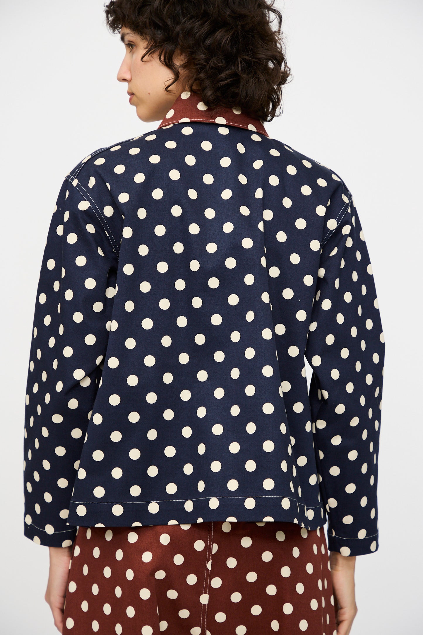 A person with short, curly hair faces away, wearing the KasMaria Cotton Twill Shirt Jacket in Polka Dot and a brown skirt with large white polka dots and patch pockets.