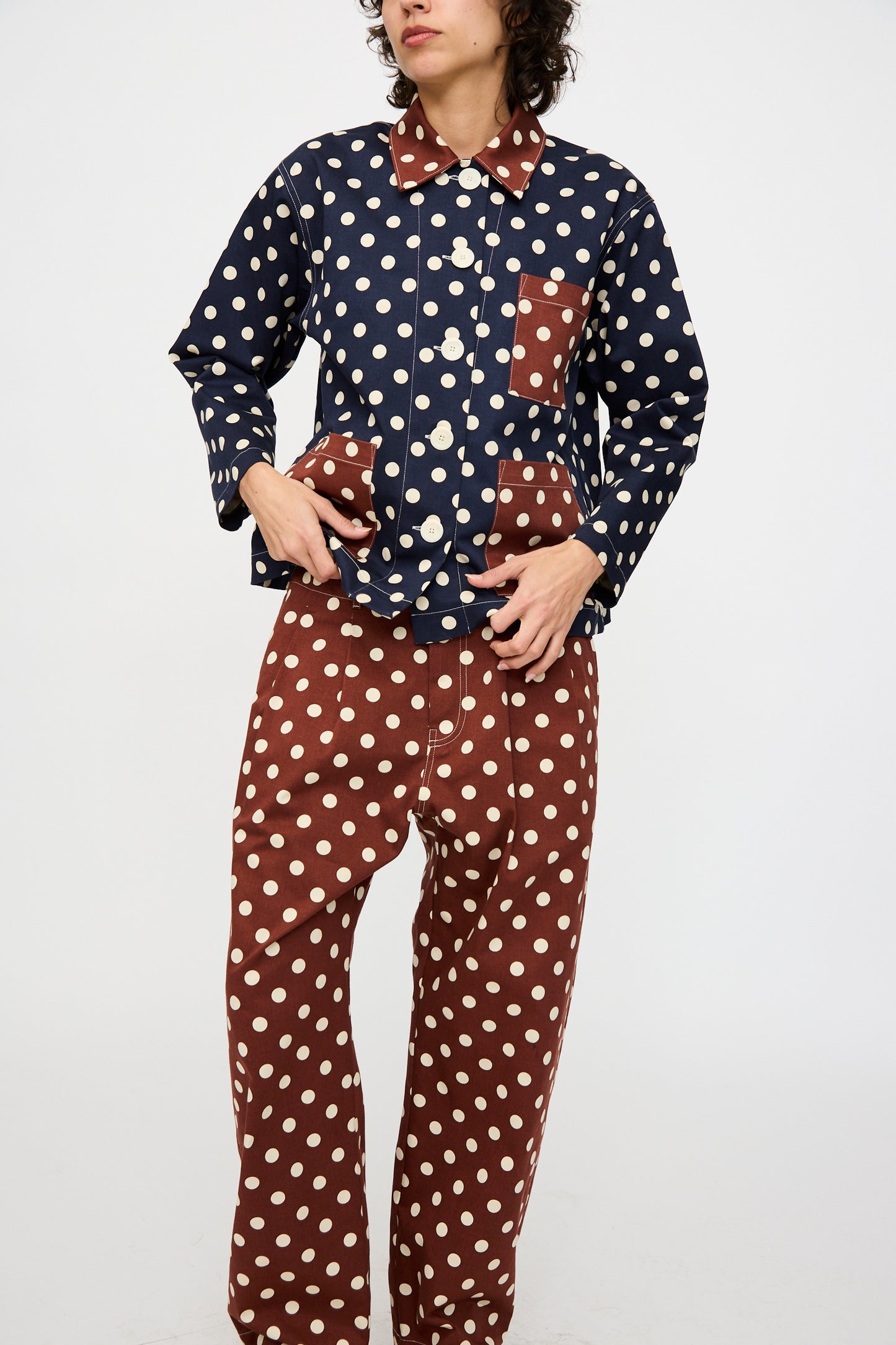 A person stands against a plain background wearing KasMaria's Cotton Twill Shirt Jacket in Polka Dot with matching brown pants, both featuring white dots and contrasting patch pockets.