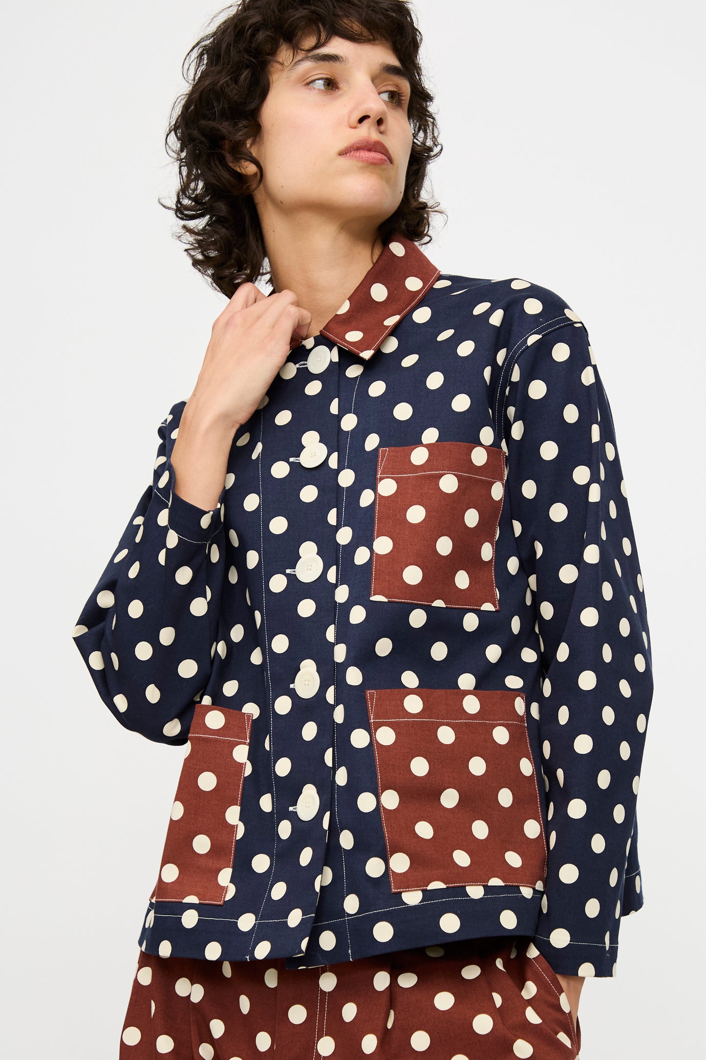 A person wears the KasMaria Cotton Twill Shirt Jacket in Polka Dot, featuring white polka dots and contrasting brown patch pockets and collar, looking to the side.