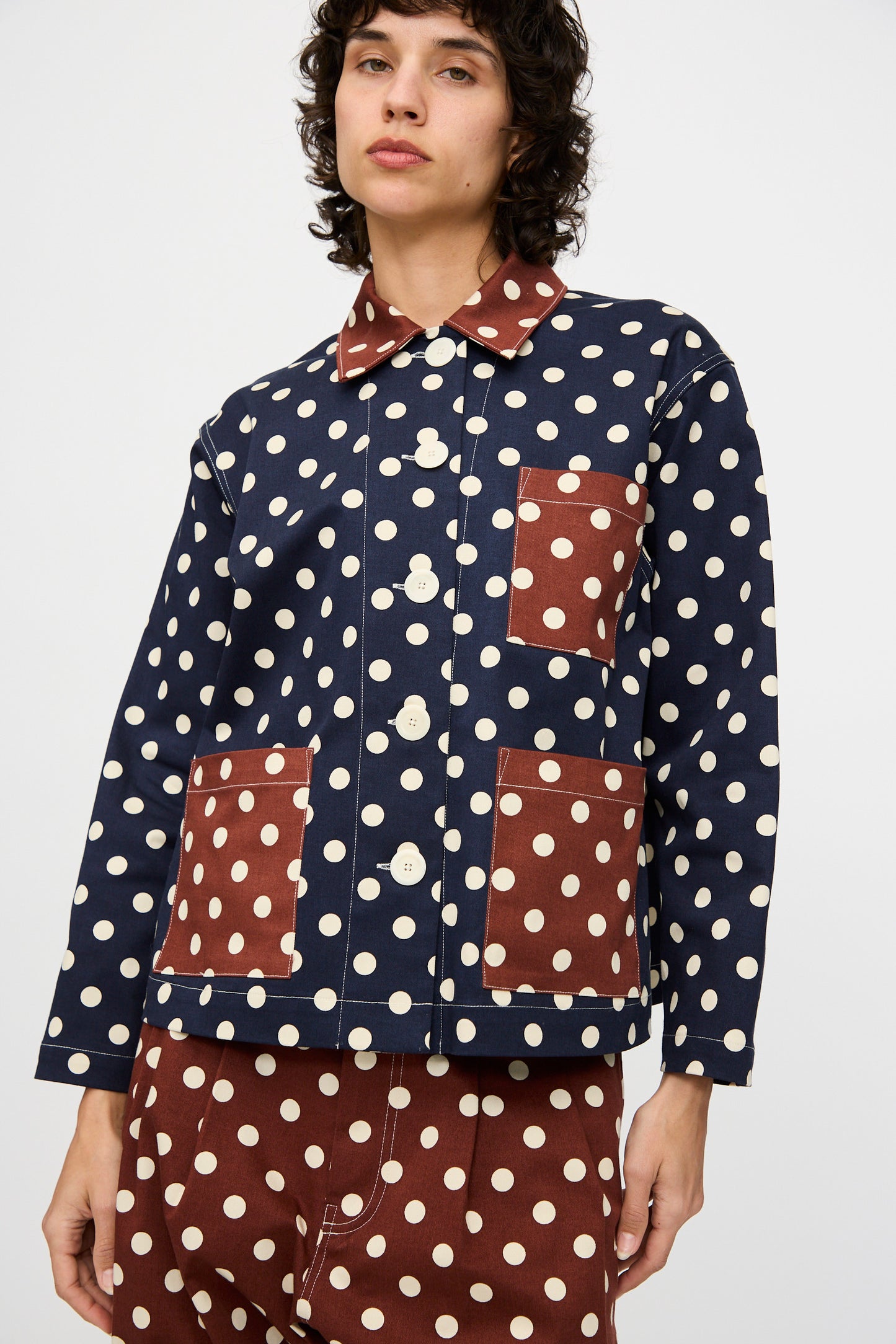 A person wears the KasMaria Cotton Twill Shirt Jacket in Polka Dot, featuring navy blue and white fabric with brown patch pockets and collar, paired with matching brown polka dot pants.