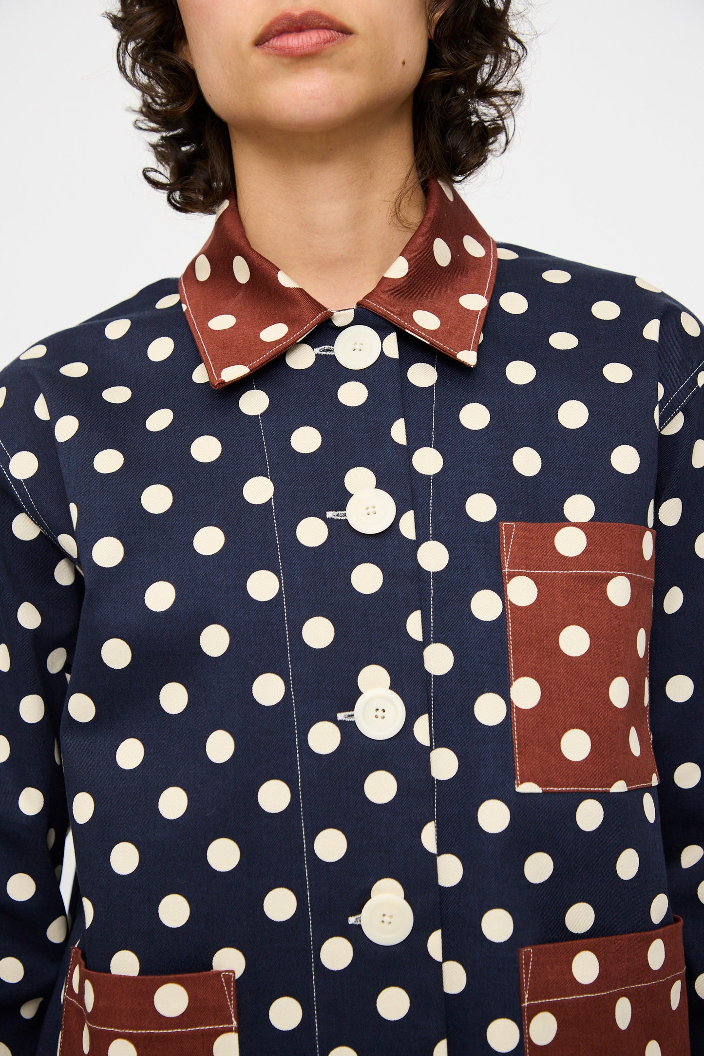 A person is shown from shoulders to mid-chest wearing the KasMaria Cotton Twill Shirt Jacket in Polka Dot—navy blue with white dots, and featuring brown polka dot patch pockets and collar.