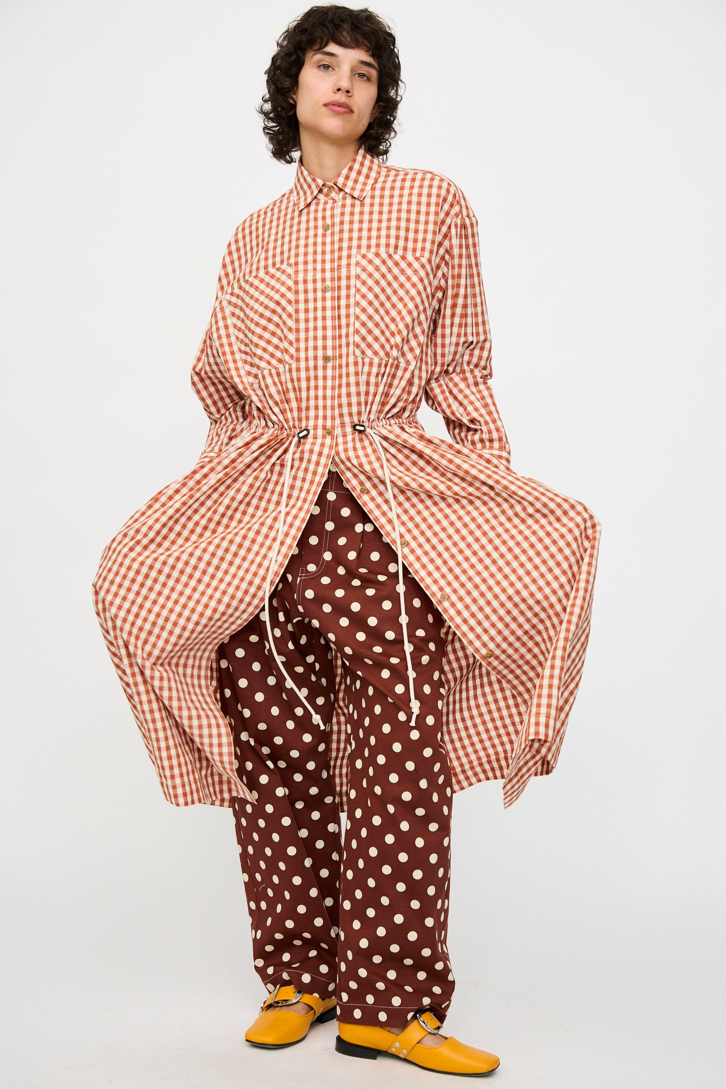 A model wears the KasMaria Crinkle Cotton Linen Drawstring Dress in Gingham over brown polka dot pants and yellow shoes, standing against a plain white background.