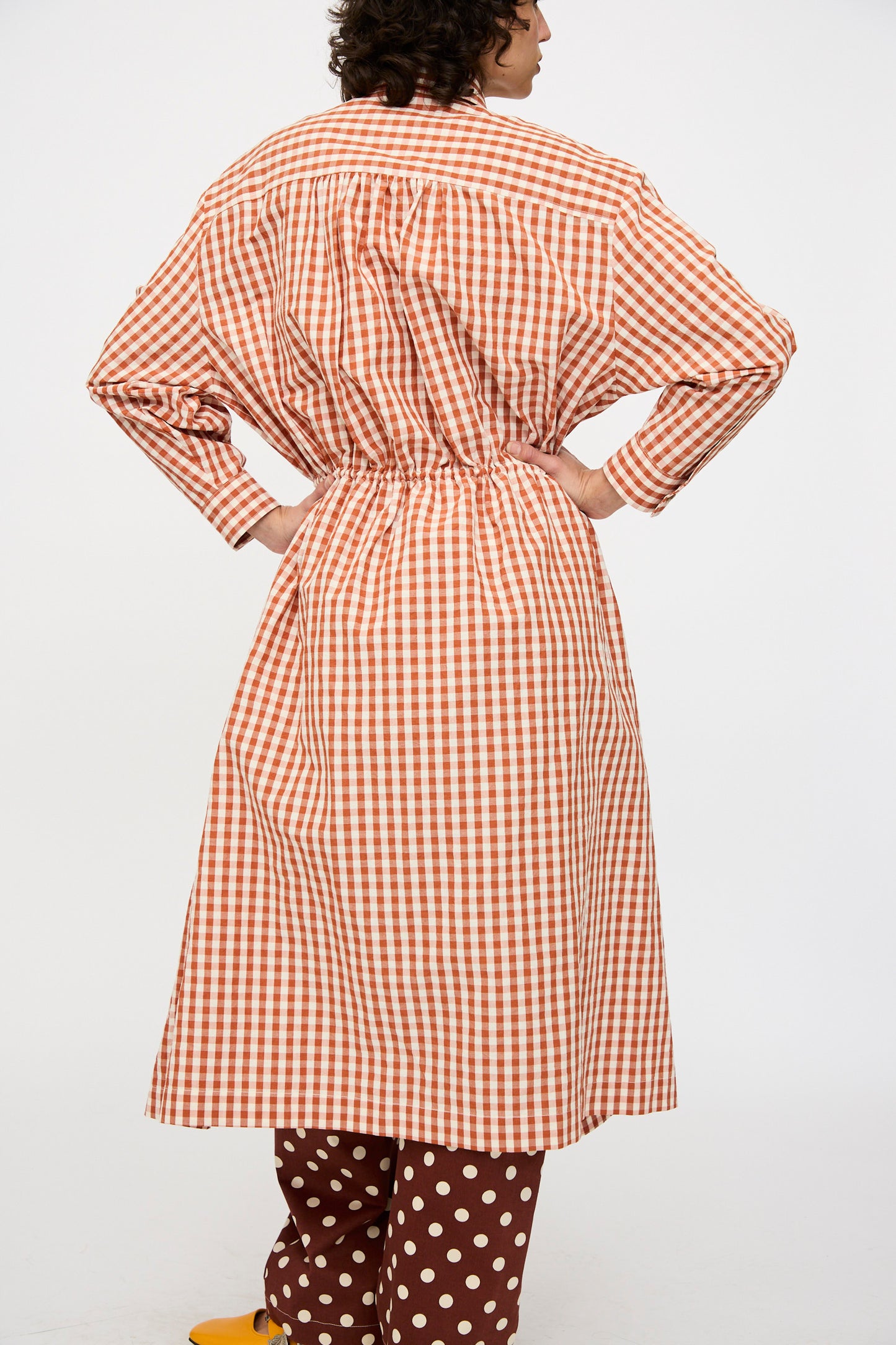 A person stands with hands on hips, viewed from the back, wearing the KasMaria Crinkle Cotton Linen Drawstring Dress in Gingham over brown polka dot pants and yellow shoes.