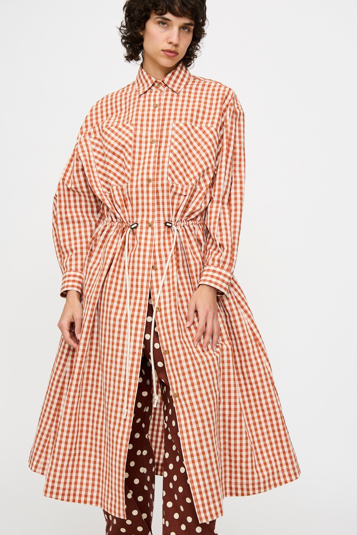 A person wearing the KasMaria Crinkle Cotton Linen Drawstring Dress in red and white gingham over brown polka-dot pants, standing against a plain background.