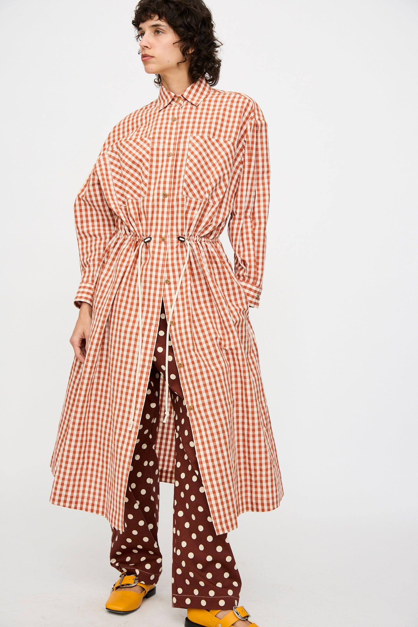 A person wearing the KasMaria Crinkle Cotton Linen Drawstring Dress in red and white gingham, paired with brown polka dot pants and yellow shoes, stands against a plain background.