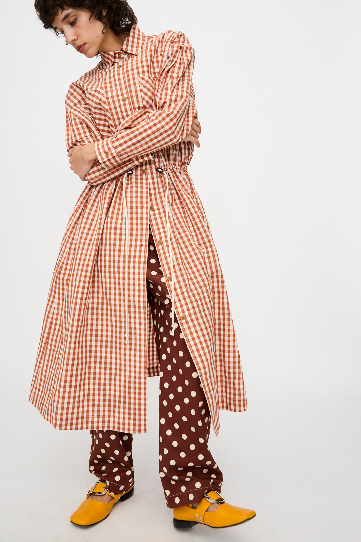 A person wearing the KasMaria Crinkle Cotton Linen Drawstring Dress in red and white gingham stands with arms crossed against a plain background, styled over brown polka dot pants and yellow shoes.