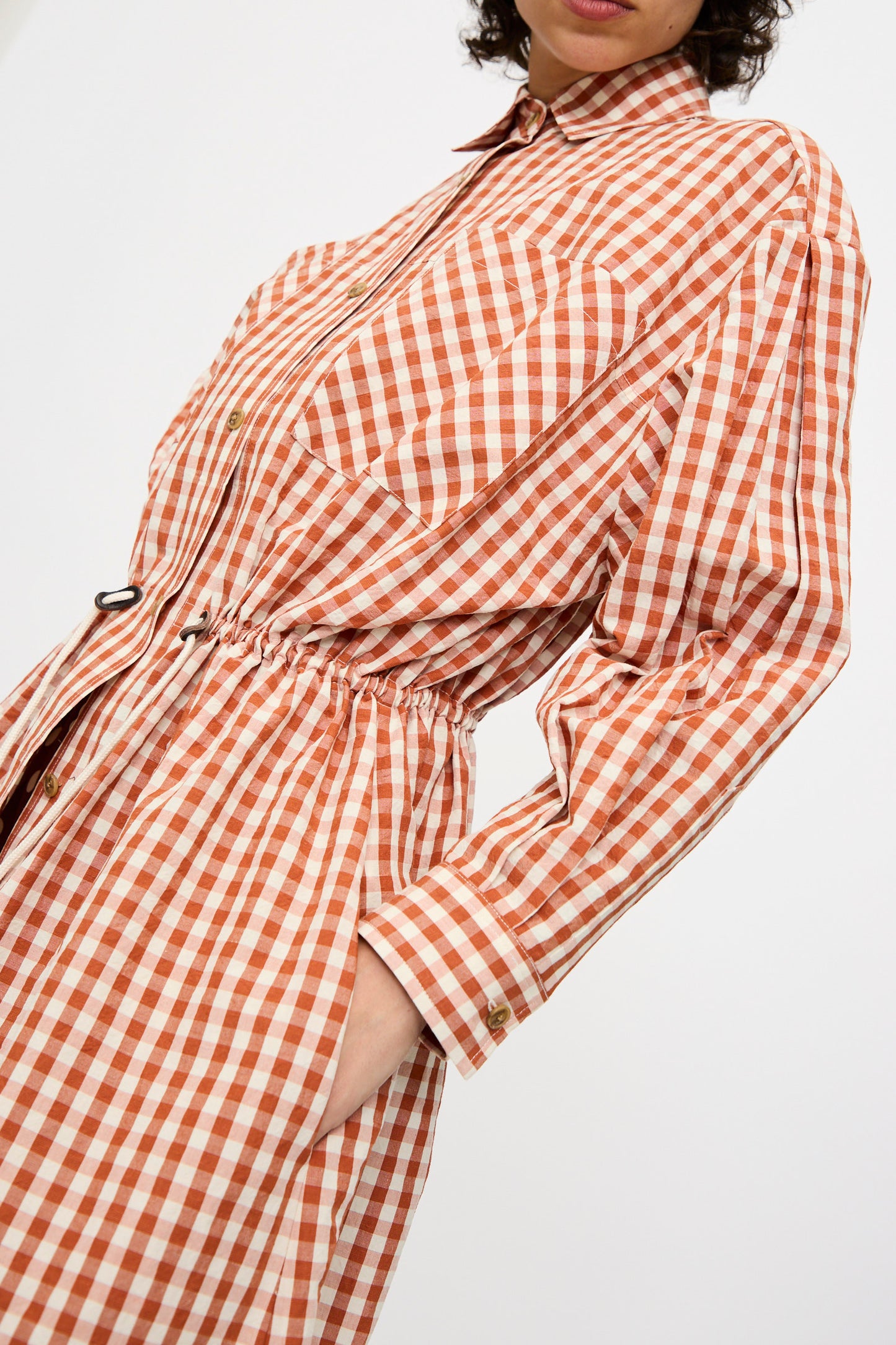 Model wears the KasMaria Crinkle Cotton Linen Drawstring Dress in Gingham, featuring red and white check pattern, long sleeves, button-up front, elastic waist, and soft cotton-linen blend. Shown from mid-face to thigh.