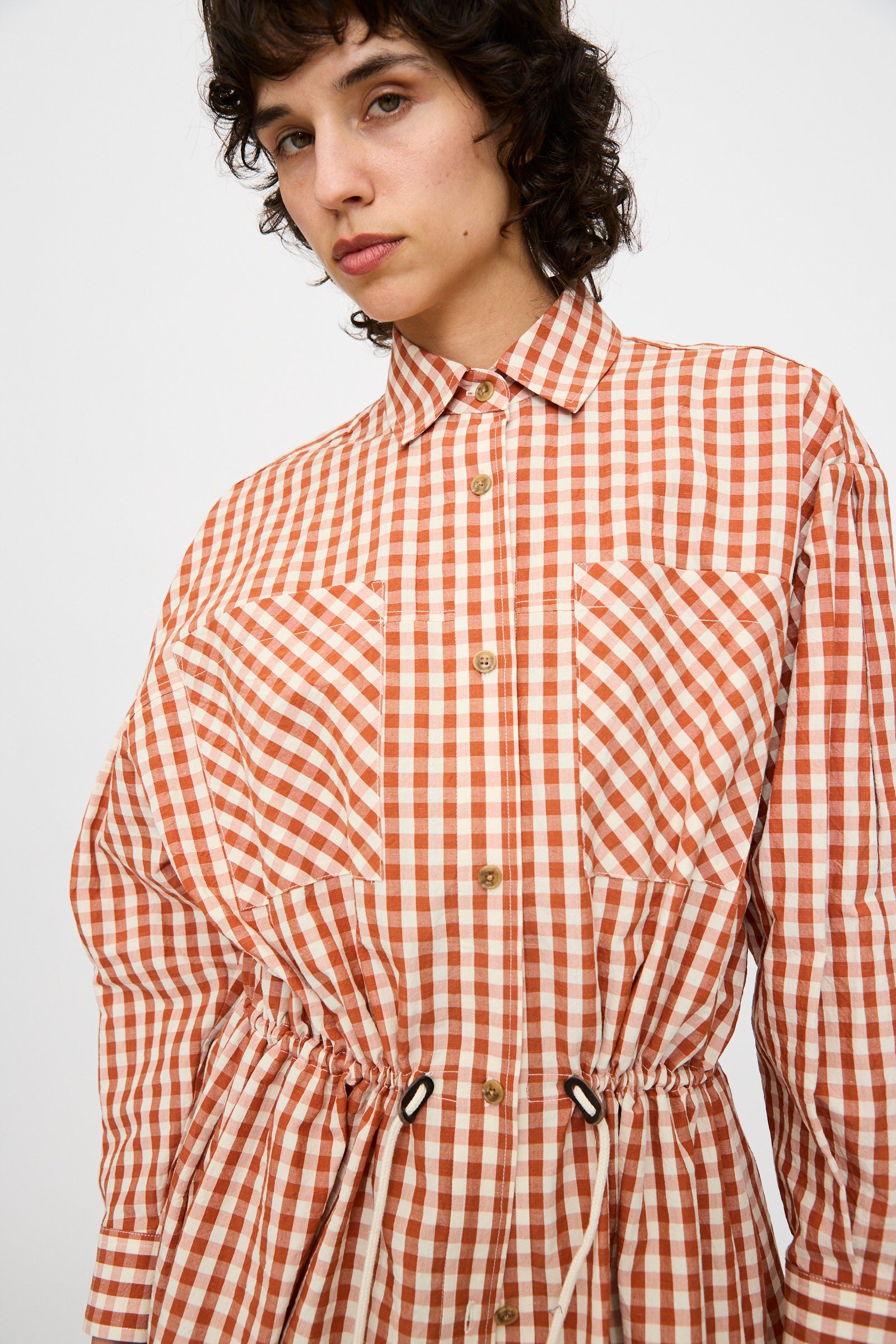 Model wears the KasMaria Crinkle Cotton Linen Drawstring Dress in orange and white gingham, featuring long sleeves, button-up front, front pockets, and a drawstring waist; made from a cotton-linen blend.