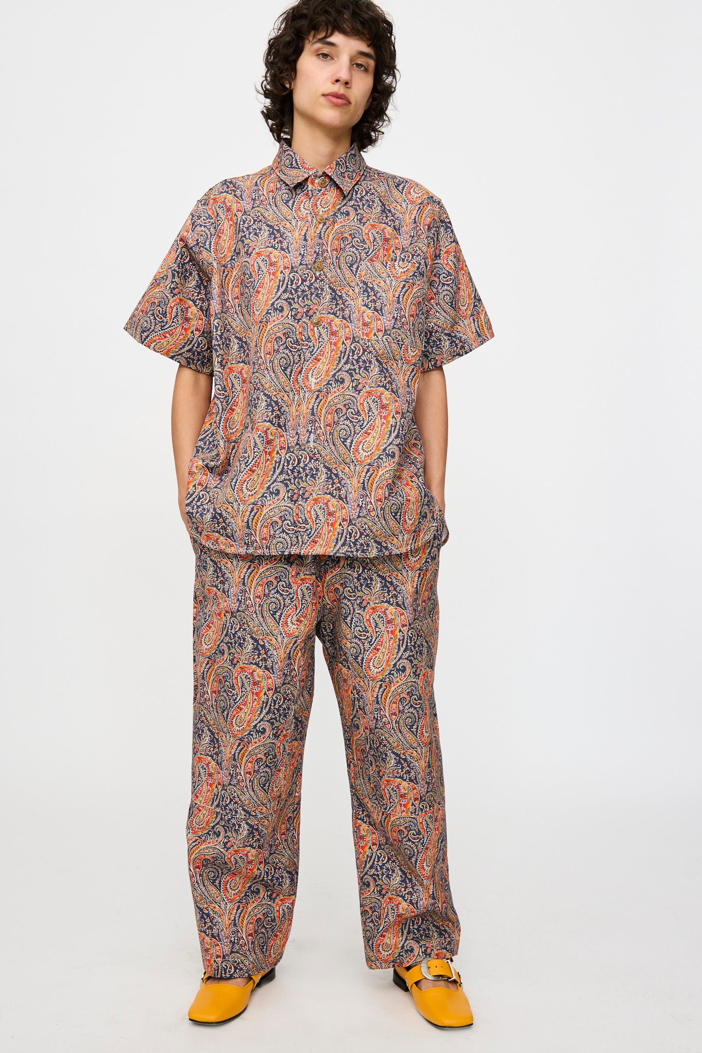 A person stands against a plain background wearing the KasMaria Liberty Cotton Canvas Shirt in Paisley with matching pants, made in NYC, paired with bright yellow shoes.