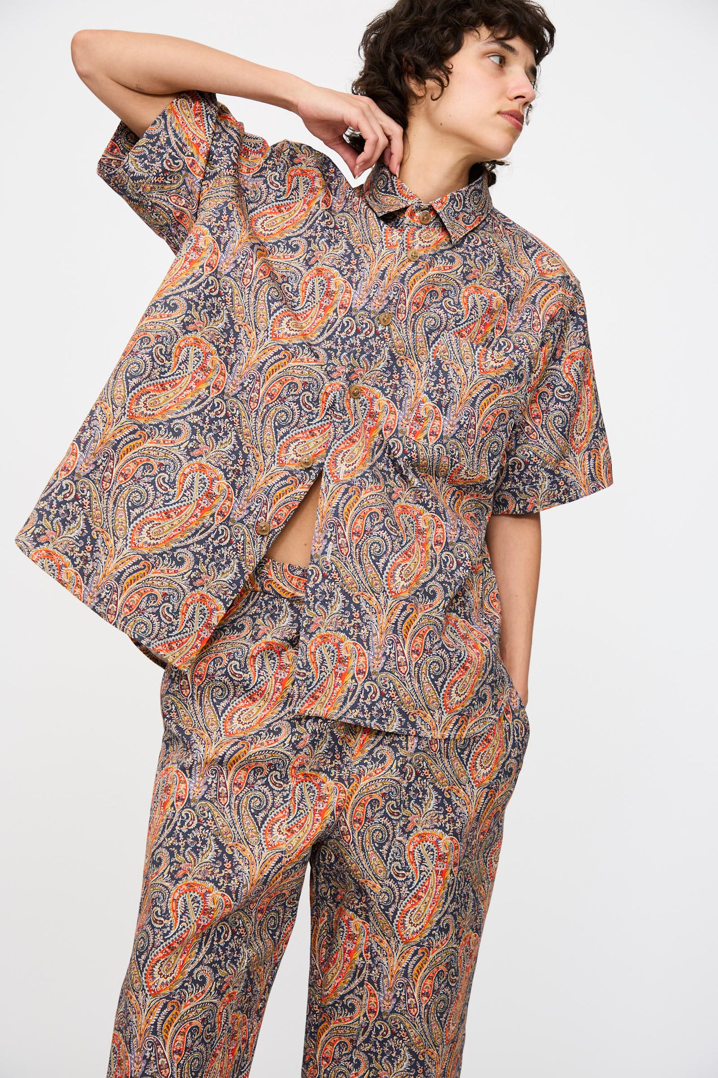 A person wearing the KasMaria Liberty Cotton Canvas Shirt in Paisley stands against a plain white background, paired with matching orange and blue pants.