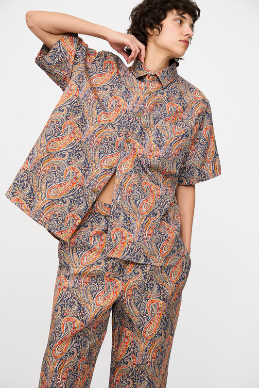 A person wearing the KasMaria Liberty Cotton Canvas Shirt in Paisley stands against a plain white background, paired with matching orange and blue pants.