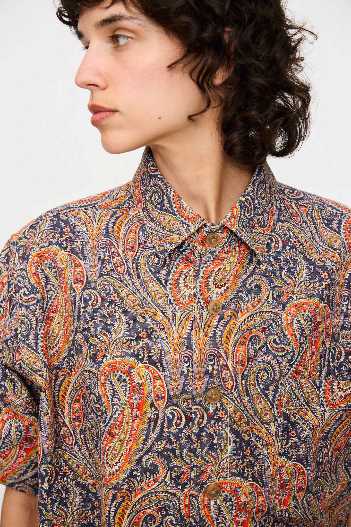 Person with short curly hair, wearing the KasMaria Liberty Cotton Canvas Shirt in Paisley, made in NYC, looks to their left against a plain background.