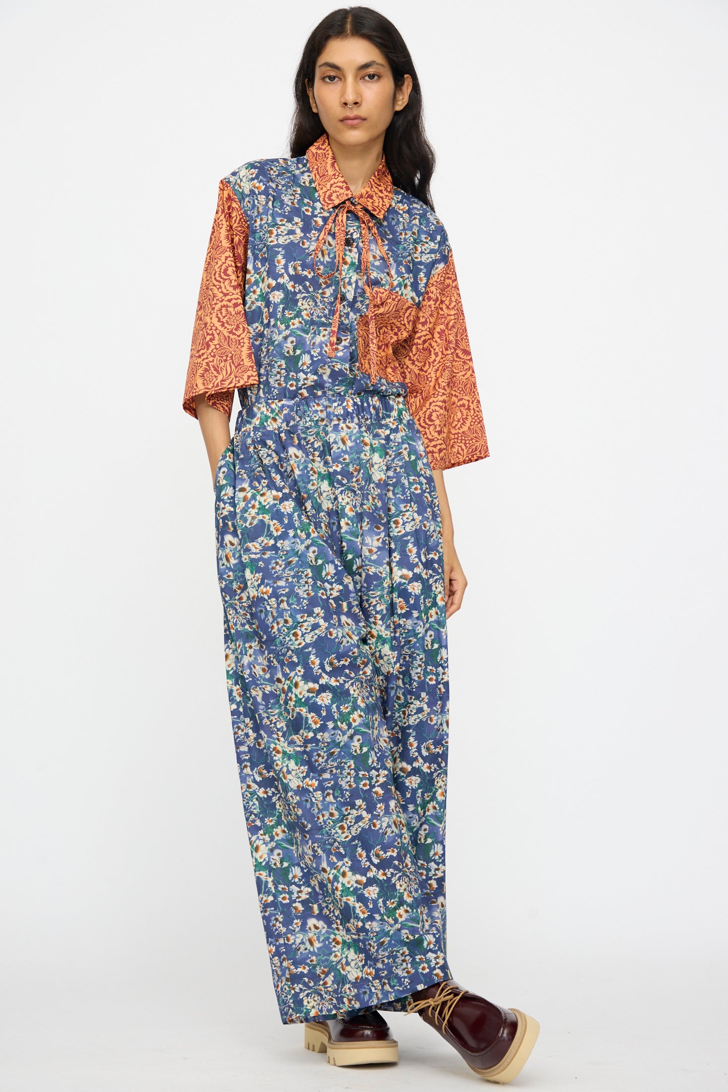 Person wearing KasMaria's Liberty Floral Baggy Pleated Trouser in Blue, paired with an orange patterned short-sleeve top and dark shoes with light soles, standing against a plain background.