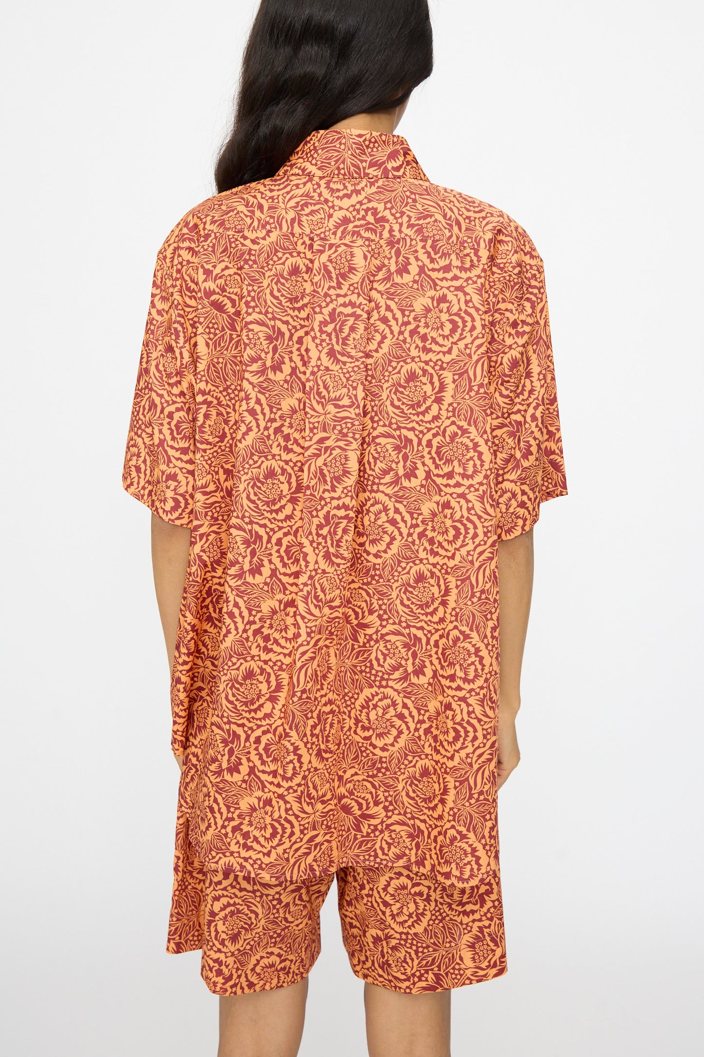 A person with long dark hair faces away, wearing the KasMaria Liberty Floral Classic Oversized Shirt in Orange, featuring a red and orange floral pattern on Liberty of London Tana Lawn Cotton, set against a plain background.