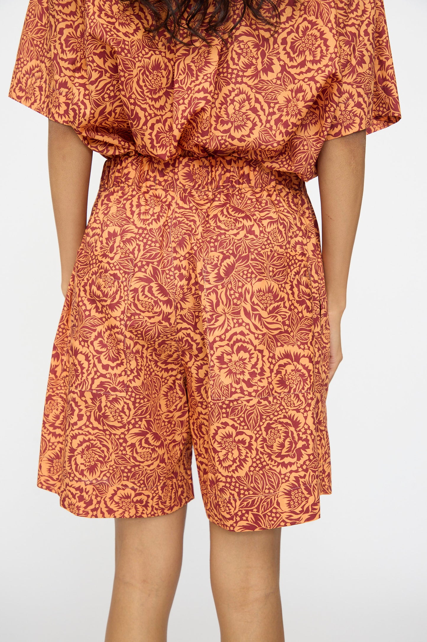 Model wears KasMaria's Liberty Floral Shorts in Orange, featuring a matching top with an orange and red floral pattern, shown from the back against a plain light background.