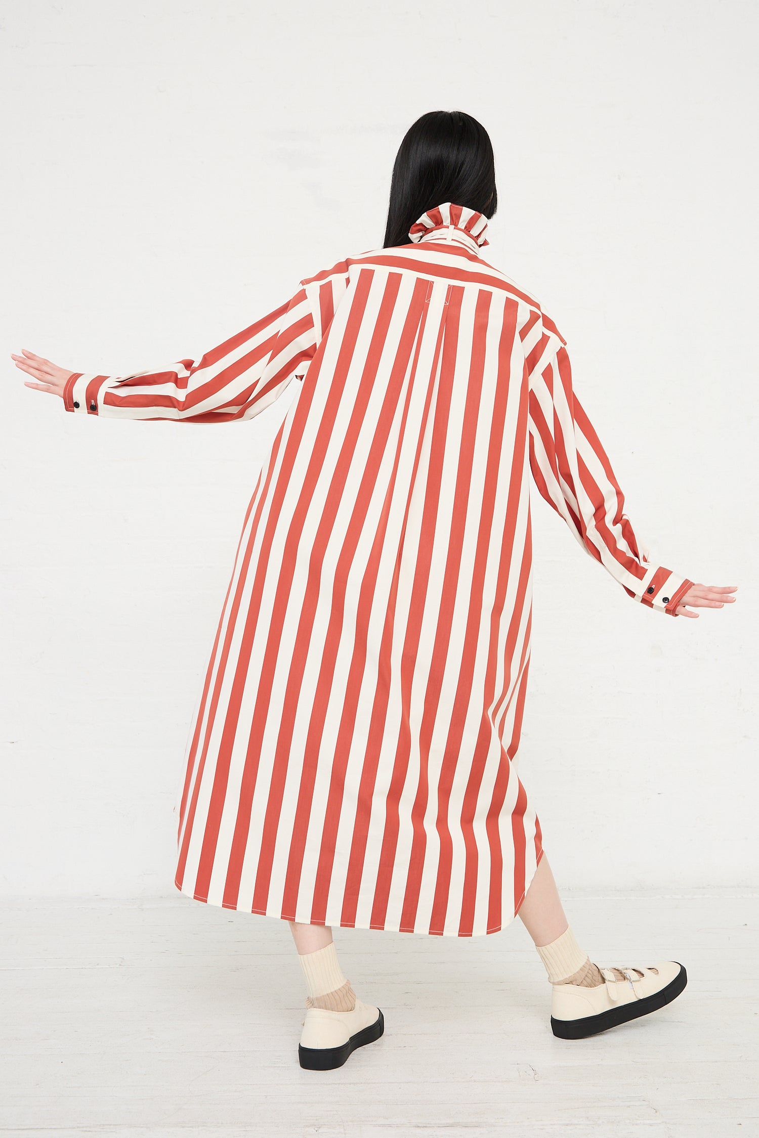 A person wearing a KasMaria Cotton Poplin Ruffle Neck Shirt Dress in Stripe with arms outstretched, viewed from behind.