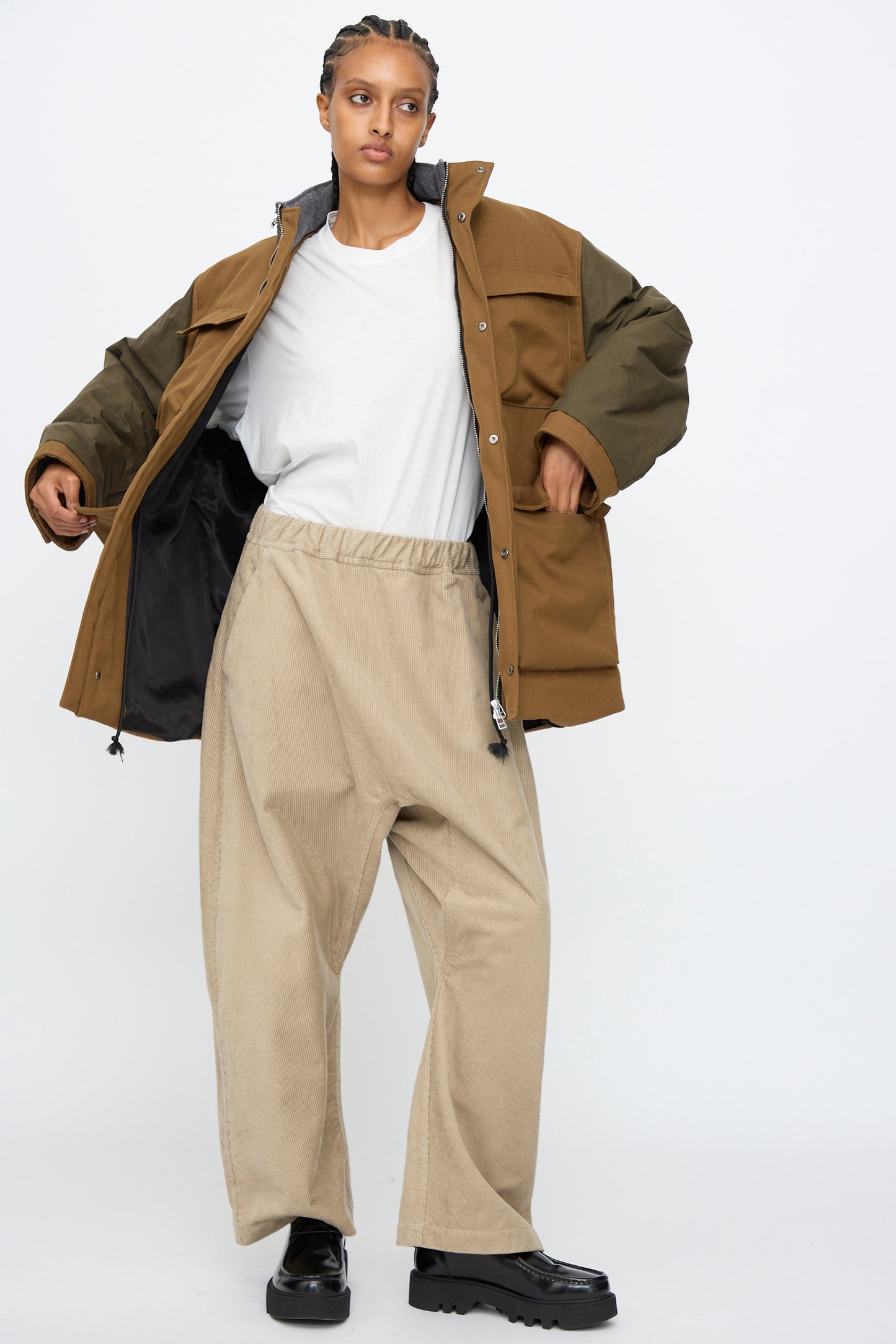 A person wears the Kassl Canvas Wax Field Jacket in Almond and Oil Black over a white shirt with beige wide-leg pants and black shoes, standing against a plain white background.
