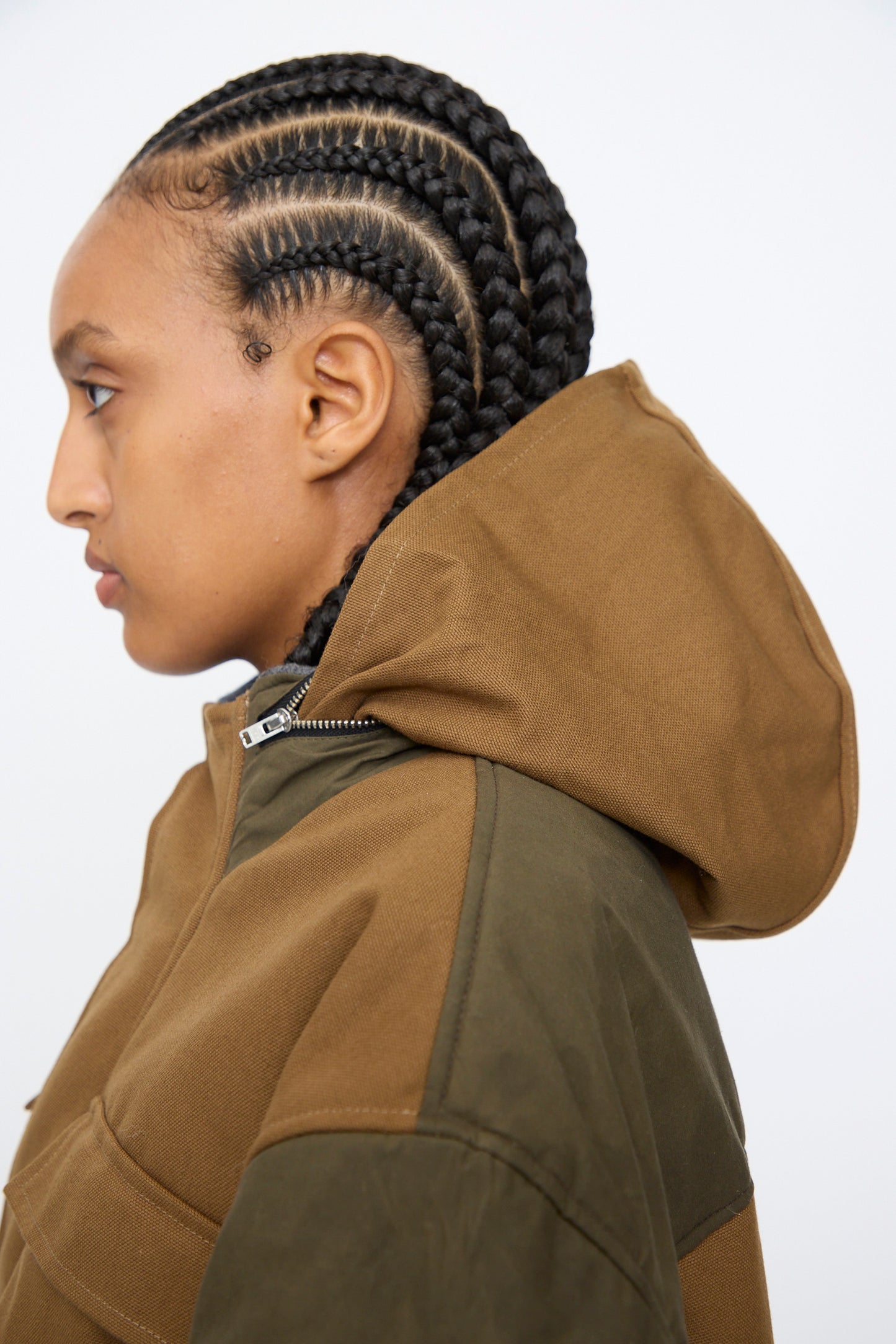 A person with cornrow braids is shown in profile, wearing the Kassl Canvas Wax Field Jacket in Almond and Oil Black, featuring an adjustable waist, against a plain background.