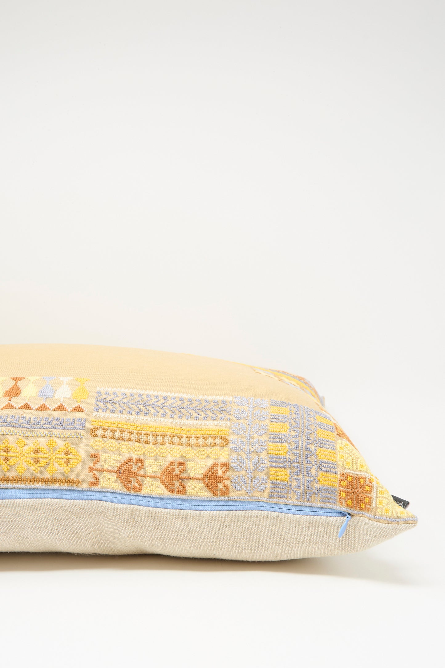 The Imm Bushra Hand Embroidered Pillow in Mustard by Kissweh features Belgian linen, a beige base, and hand-embroidered yellow, orange, and blue geometric patterns, finished with a light blue edge zipper.