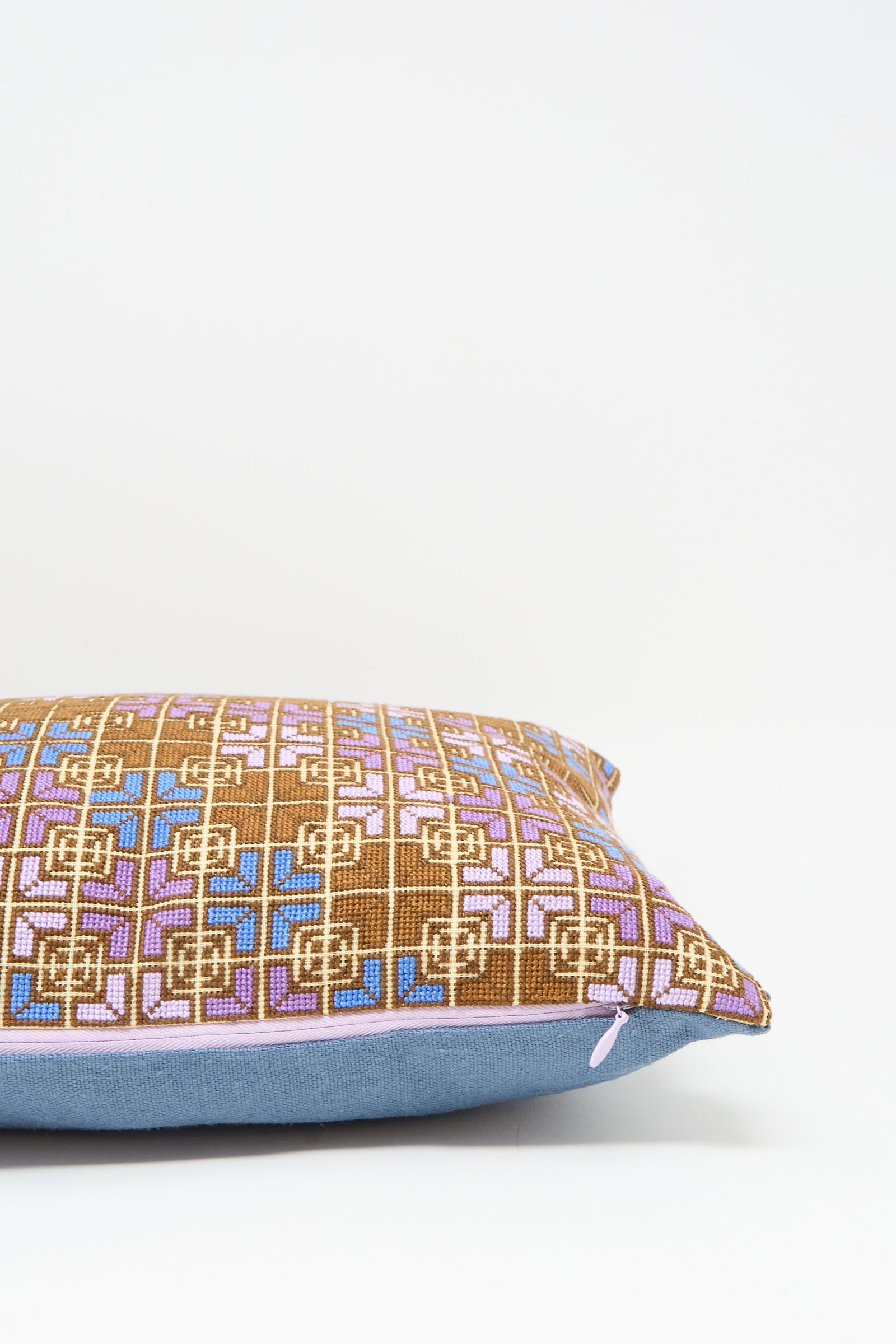 The Kissweh Rana Hand Embroidered Pillow in Lilac and Cognac features a geometric brown, pink, and blue pattern atop a solid light blue bottom, shown against a plain white background.