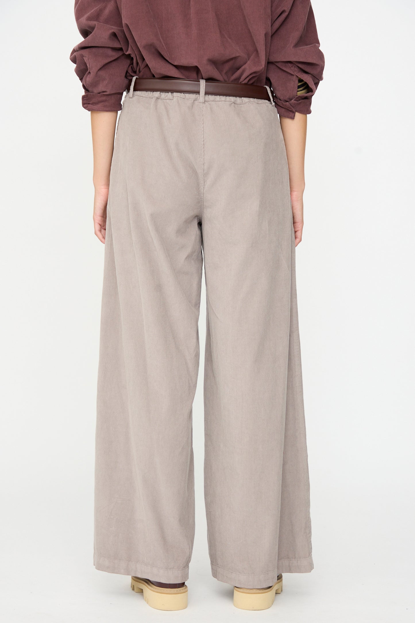 A person stands facing away against a plain white background, dressed in a brown long-sleeve shirt and Labo.Art’s Binocchio Trouser in Malva—beige wide-leg pants crafted from soft cotton needlecord—paired with a brown belt and beige shoes.
